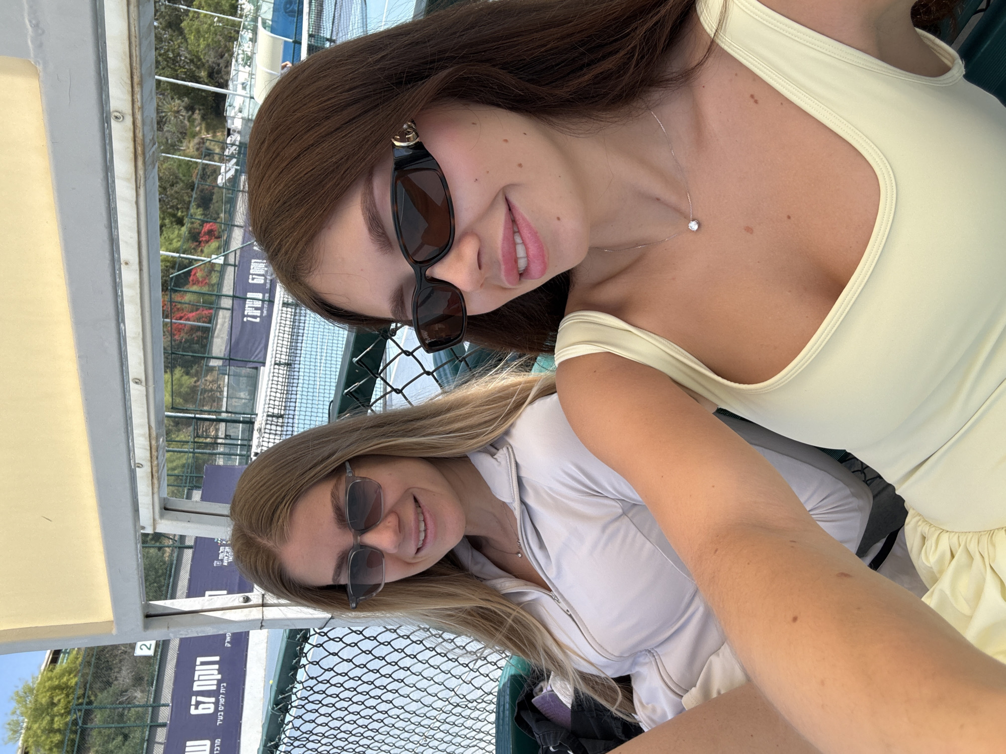 Two women are sitting outside on a sunny day, with a tennis court and fence visible in the background. The woman on the right has long brown hair, is wearing sunglasses and a light yellow top, and a necklace. The woman on the left has blonde hair, is also wearing sunglasses, and a white jacket. There is a sign in Hebrew behind them. The location is Tel-Aviv, Israel, as per the sign.