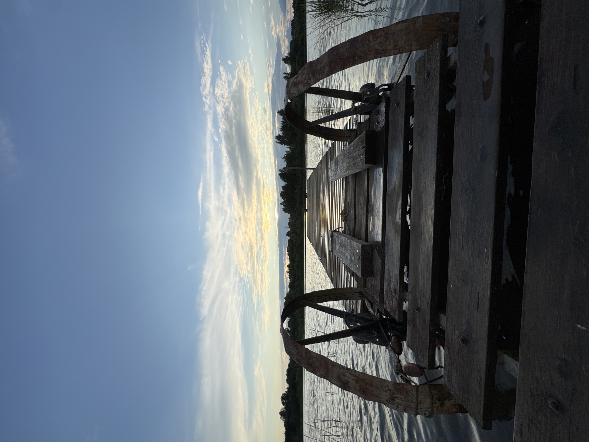 The image shows a wooden pier extending over a body of water. The pier is weathered and appears to be made of dark planks. On either side of the pier, towards the foreground, are two rusted, curved metal structures, possibly part of a winch or mooring system. The water is calm with gentle ripples. In the background, a line of trees borders the water under a sky with scattered clouds, tinged with the colors of sunset. The overall scene suggests a natural, tranquil environment, likely a lake or slow-moving river, possibly in or near Sitno, Russia, given the vegetation and water. There are no people or animals visible in the image.