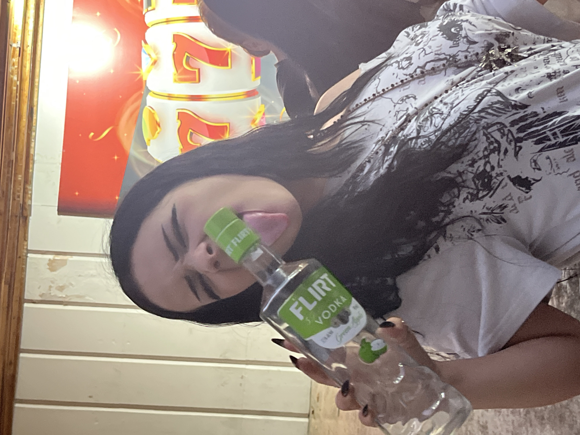 A young woman with long dark hair and heavy eyeliner holds a small bottle of "FLIRT VODKA" with a green label and its cap off, its opening pressed against her tongue. She is wearing a white t-shirt with a black, intricate graphic print and a beaded necklace with a large pendant. The background shows part of a slot machine with three "7" symbols illuminated in red and yellow, suggesting a casino or gambling establishment. The wall behind her appears to be paneled with vertical white planks, some showing peeling paint. The image is an outdoor shot, possibly in a public space in Vratsa, Bulgaria, given the context. The woman's pose and the open vodka bottle suggest a moment of playful or perhaps reckless behavior.