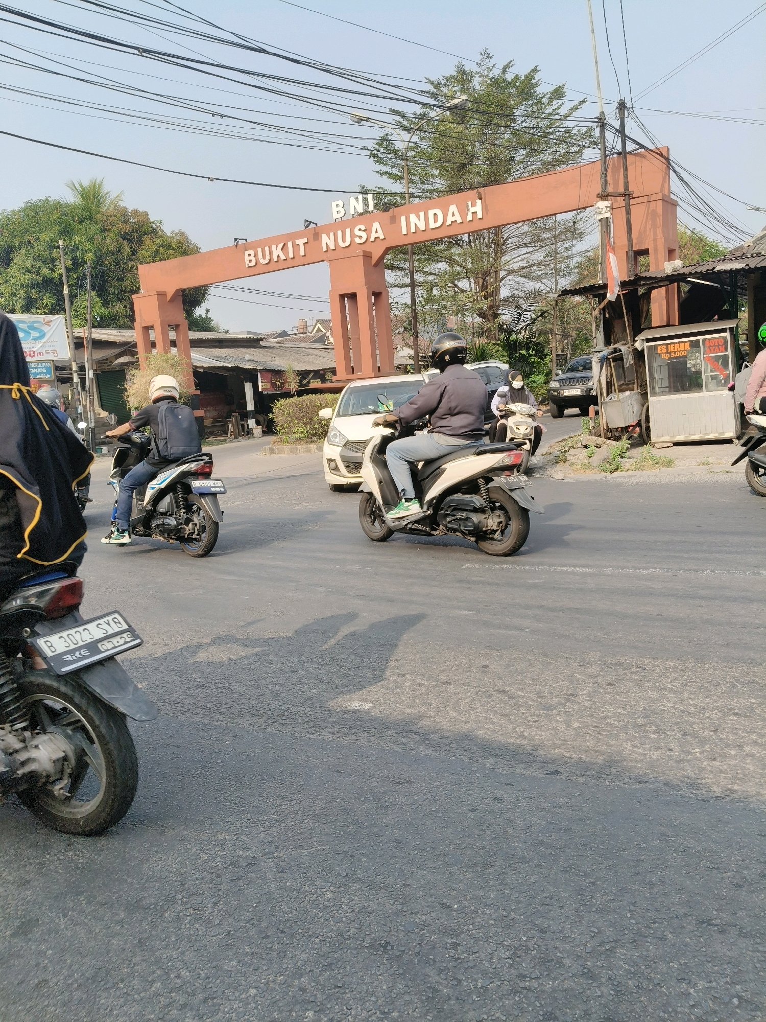 The image captures a street scene in South Tangerang, Indonesia, featuring a prominent orange archway with the text "BNI BUKIT NUSA INDAH" emblazoned across it. Several individuals are riding motorcycles, navigating the asphalt road under a clear sky with visible power lines overhead.
In the foreground, a black motorcycle is parked, with its license plate partially visible. To the left, a person wearing a black and yellow garment is partially visible. Further back, a person in a gray jacket and blue jeans rides a white scooter, carrying a dark backpack. Another person on a white scooter is seen in the background.
The environment includes a white car, a black SUV, and several small structures and shops lining the roadside. One stall displays "ES JERUK Rp 5.000" and "SOTO AYAM," suggesting a food and beverage vendor. Trees and other greenery are present, contributing to the urban landscape. The overall impression is a typical street view in a populated area, possibly a commercial or residential district within South Tangerang.