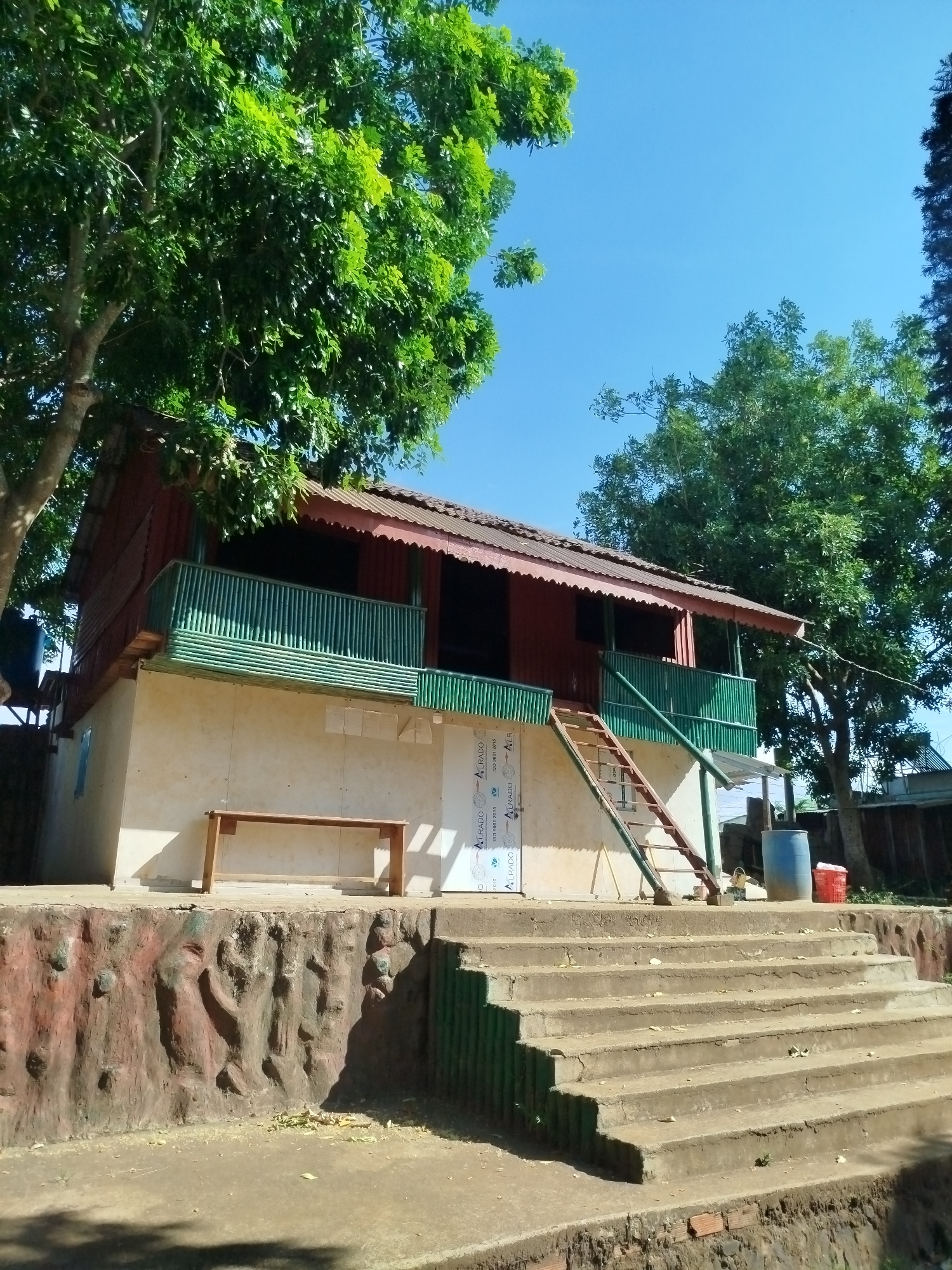 Here is a forensic description of the image:
The image depicts a two-story building situated in an outdoor environment. The building has a red roof and upper walls, while the lower walls are a lighter color. A green railing surrounds the upper level. A ladder leads to the upper level.
A set of concrete steps leads up to the building's entrance area. A low, decorative wall with a textured surface borders the area in front of the steps. A wooden bench sits on the lower level in front of the light-colored wall.
A water tank is visible near the building's left side. A large tree with dense green foliage partially obscures the building.
The image was taken in An Phu Ward, Vietnam.