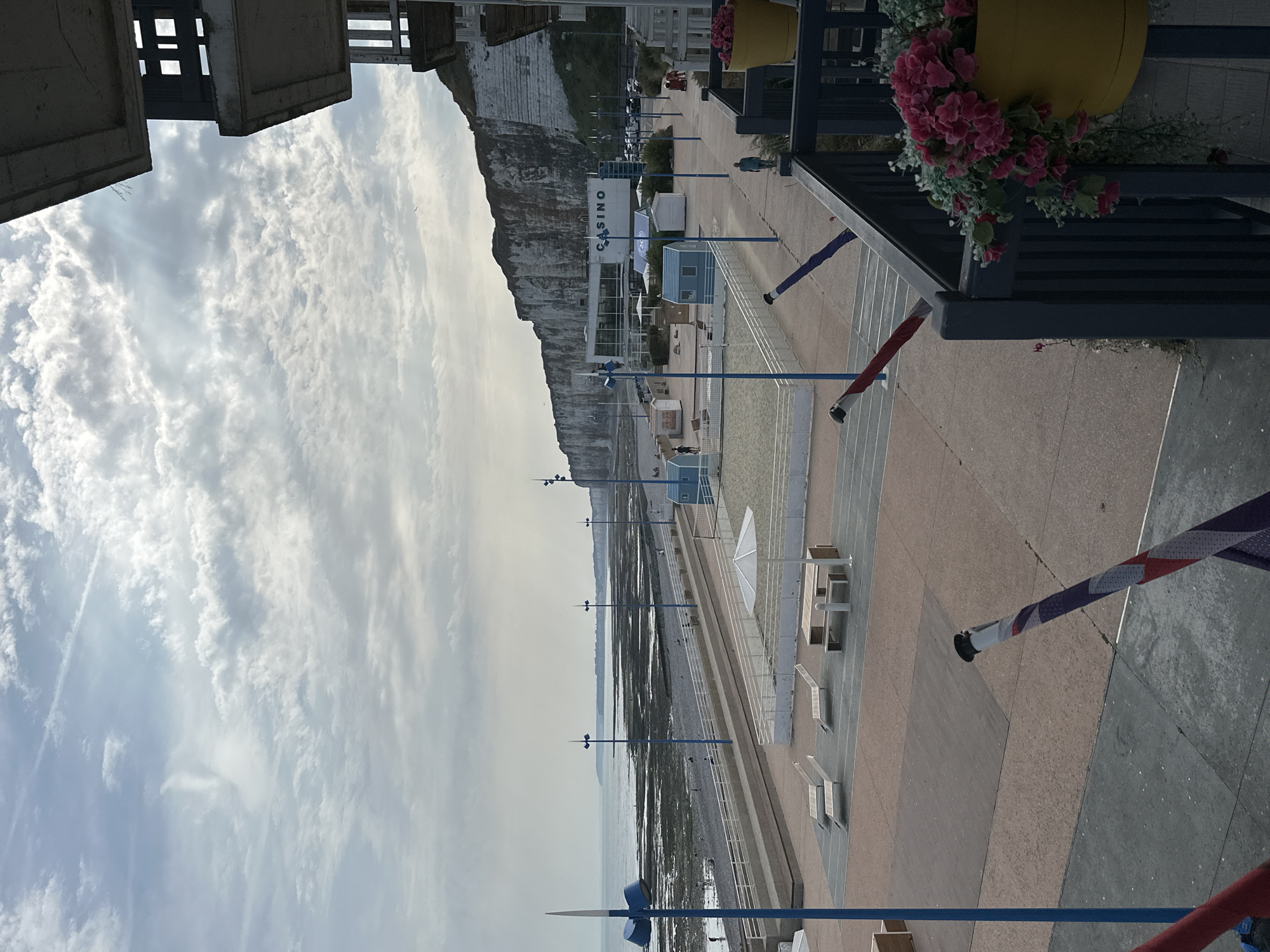 The image depicts a seaside promenade in Saint-Valery-en-Caux, France, featuring a beach, a casino building, and apartment complexes. The sky is overcast with scattered clouds, and the sea is visible in the background. On the promenade, there are lounge chairs, picnic tables, and a sandy area with a volleyball net. A blue beach hut and a white umbrella are also present.
To the right, a portion of a dark blue balcony railing is visible, adorned with a yellow planter containing pink flowers. The casino building is situated in the middle ground, its name clearly displayed in large white letters. Behind the casino, steep white cliffs rise up, characteristic of the region. Several cars are parked in front of the casino, and a few people are walking on the promenade and the beach. The overall atmosphere is calm and appears to be during daylight hours.