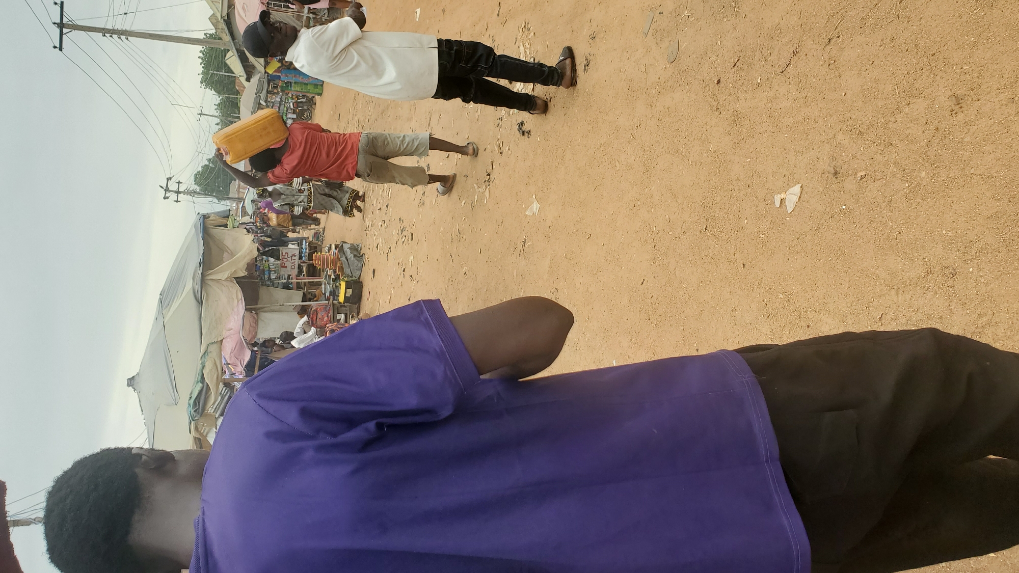 In the city of Jalingo, Nigeria, an outdoor marketplace scene unfolds under an overcast sky. In the foreground, a person wearing a purple t-shirt and black pants is captured from behind, their shoulder and arm visible. Further into the scene, another individual in a red shirt and light-colored shorts carries a large yellow jerrycan on their head, walking away from the camera. To their right, a person in a white short-sleeved shirt and black pants, wearing a black baseball cap, faces the camera. In the background, numerous market stalls with various coverings create a vibrant, bustling atmosphere. The ground is sandy and littered with debris. The presence of power lines overhead indicates the urban setting.