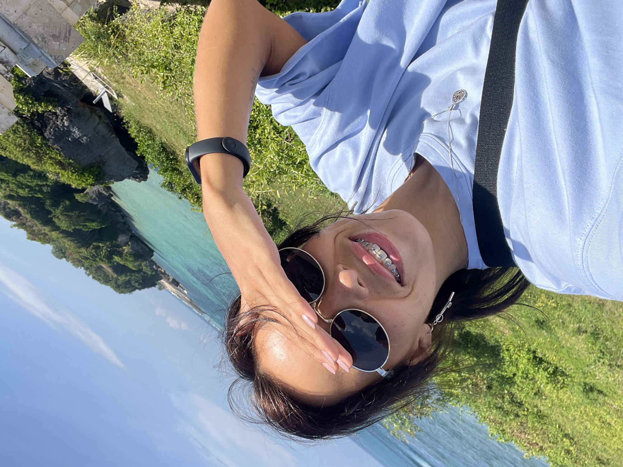A young adult female wearing sunglasses, braces, and a light blue shirt is depicted in a selfie-style photograph taken at a coastal location in Kuta Selatan, Indonesia. She shields her eyes with her right hand. The background includes a coastline with vegetation, turquoise-colored water, and a bright sky. The photograph is rotated, with the sky at the bottom and the vegetation at the top.