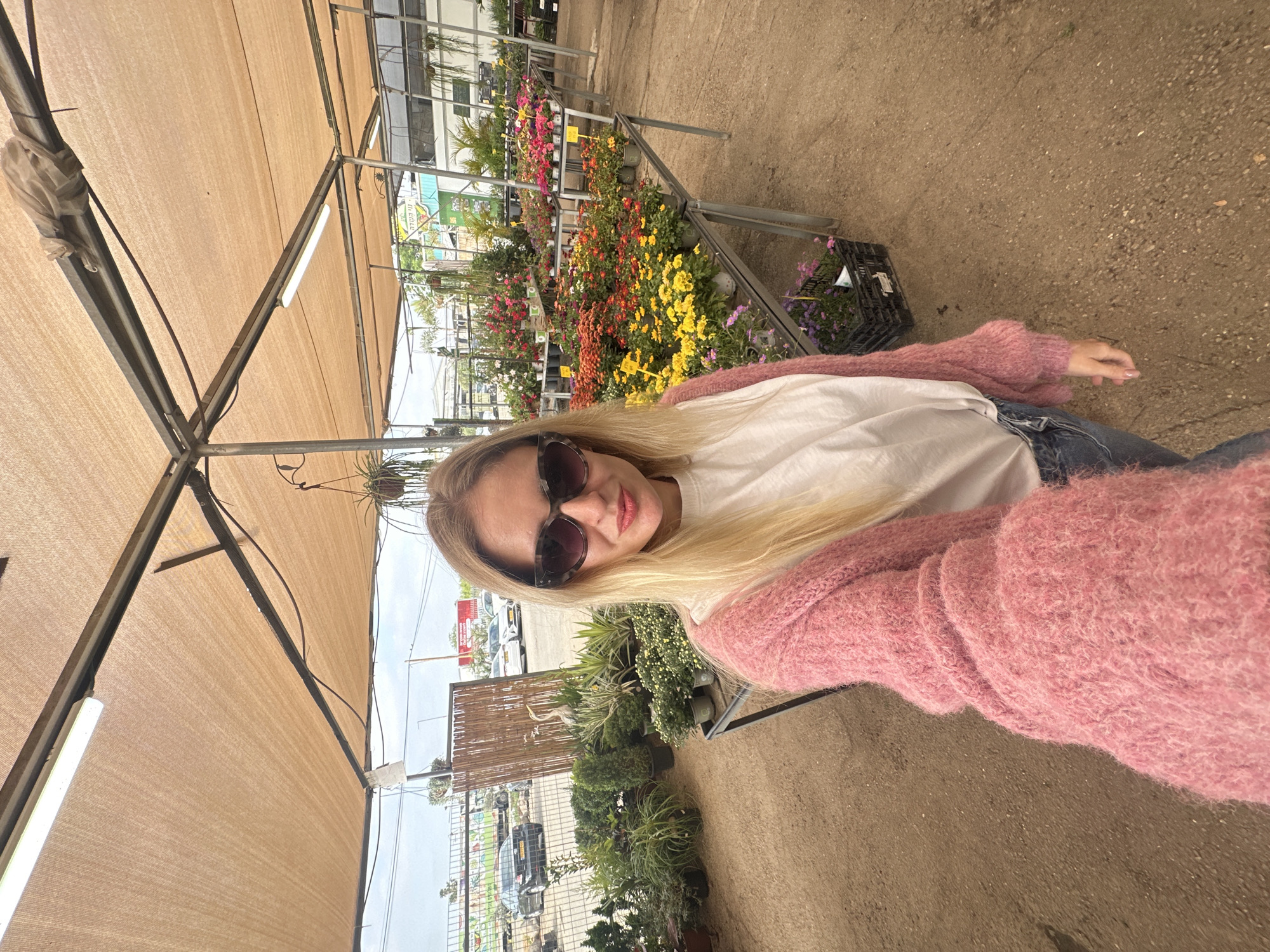 A selfie captures a woman with blonde hair, wearing dark sunglasses, a white shirt, and a pink cardigan, set in what appears to be a garden center or plant nursery.
The location is suggested to be in Petah Tikva, Israel.
The woman stands amid rows of various plants and flowers displayed on metal shelving, typical of a nursery setting. The ground is paved with light-brown concrete. Above, there is a sunshade structure with lights, providing shade over the area. In the background, through the open sides of the nursery, there is a glimpse of an outdoor environment with vehicles and possibly street signs, consistent with a city setting.