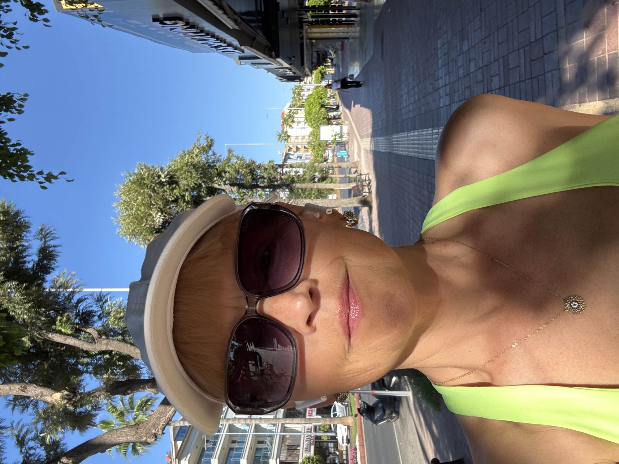 A selfie of a woman with blonde hair, wearing a visor, sunglasses, and a neon green tank top, is taken outdoors on a sunny day in Alanya, Turkey. She is looking directly at the camera. In the background, a street scene unfolds with a sidewalk, trees, buildings, and a few people walking. The woman is wearing a necklace with a circular pendant. There is a scooter parked on the left side of the image. The setting appears to be a commercial area with shops lining the street.