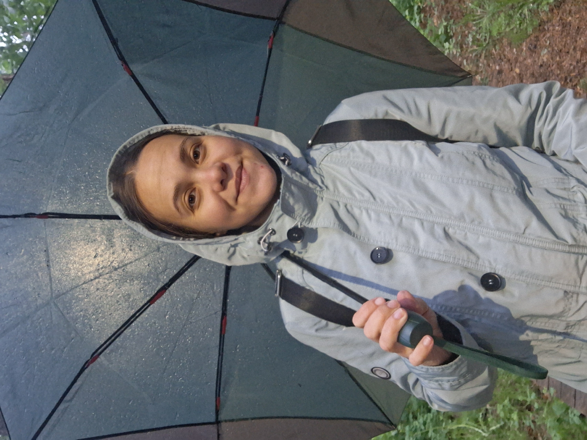 A woman stands under an open umbrella, holding it with her right hand. She is wearing a light gray hooded jacket, with the hood up. A black strap crosses her chest. The umbrella is a dark color with visible raindrops on the surface. The background suggests an outdoor location with trees and foliage. Based on the prompt, this is in Chaikovsky, Russia.