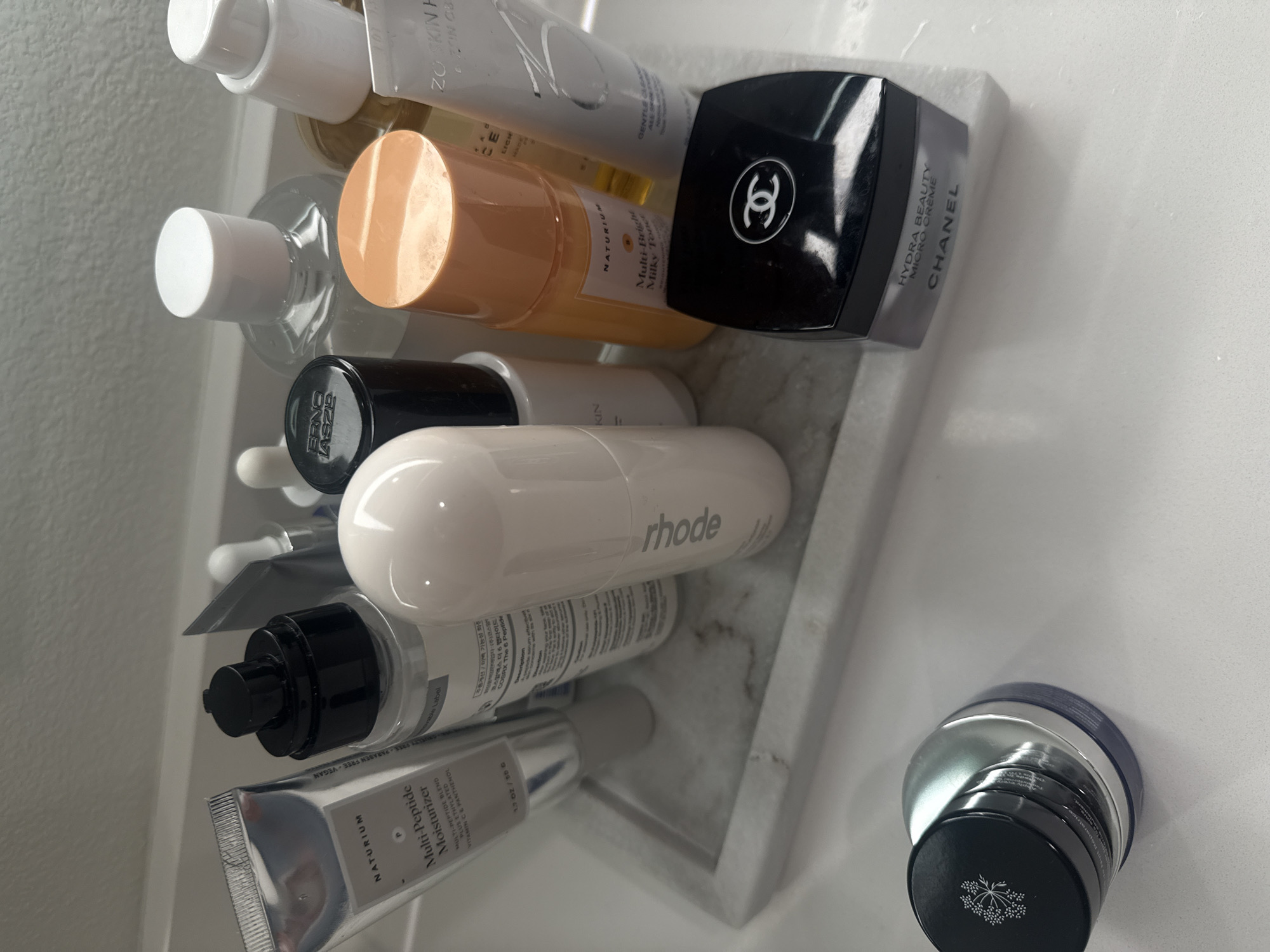This image displays a collection of various skincare products neatly arranged on a light-colored, possibly marble or stone, tray or shelf. The setting suggests a bathroom vanity or a dedicated personal care area, likely in a residential home in Los Angeles, United States of America. The lighting is bright and even, indicating an indoor environment.
Multiple distinct products are visible:
- A black and silver jar from CHANEL, prominently featuring the brand's logo and labeled "HYDRA BEAUTY MICRO CRÈME".
- An orange/peach colored bottle from NATURIUM, identified as "Multi-Bright Milky Toner".
- A white tube, partially visible, from ZO SKIN HEALTH, with "GENTLE CLEANSE" visible.
- A black bottle with "ERNO LASZLO" branding.
- A white cylindrical bottle bearing the "rhode" logo.
- A white tube, partially obscured, from COSRX, referencing "The 6 Peptide".
- A silver tube from NATURIUM, labeled "Multi-Peptide Moisturizer", with details including "MULTI-PEPTIDE BLEND PLUS ETHYLATED VITAMIN C & PANTHENOL" and "CRUELTY FREE PARABEN FREE VEGAN".
- Other generic white and clear bottles are also present, along with a small dark blue/black jar with a silver lid.
The products are arranged in an organized manner, implying a daily skincare routine. The selection includes both high-end and popular contemporary brands.