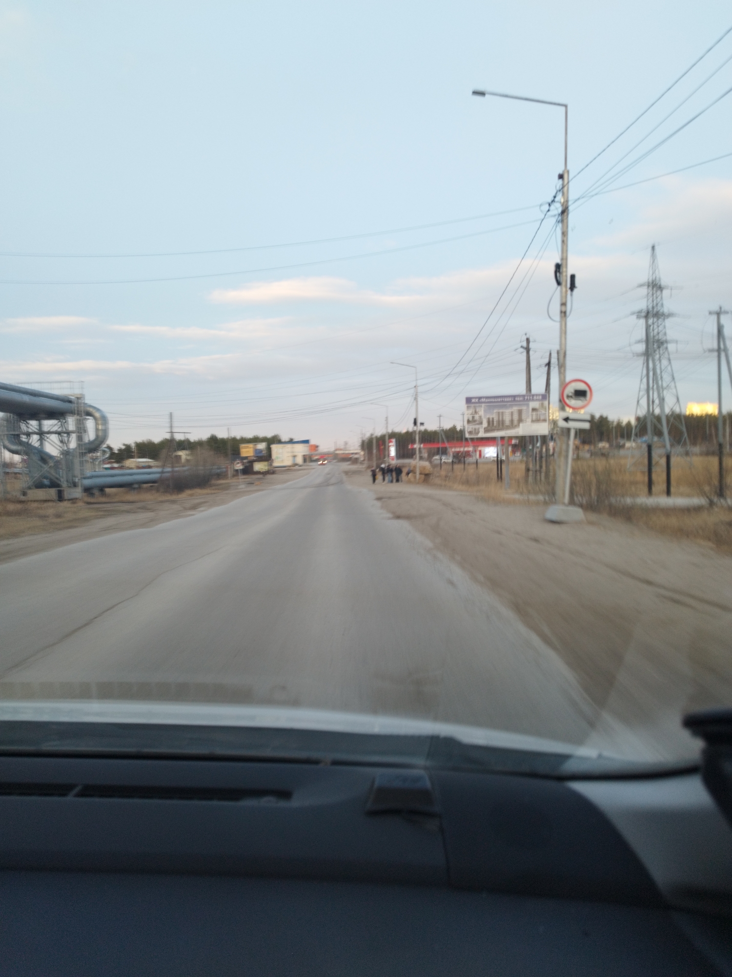 The image shows a road in a rural or semi-rural area, likely on the outskirts of a city. The view is from inside a vehicle, looking down the road. The road appears to be paved but worn, with visible cracks and possible debris. On the left side of the road, there are large industrial pipes supported by a metal structure. On the right side, there are utility poles and an electrical transmission tower. Further down the road, there are several people walking. A building with signage is visible in the distance, possibly a store or business. A traffic sign restricting trucks is also visible on the right. The sky is overcast with hints of sunlight breaking through, suggesting either early morning or late afternoon. Based on the style of the buildings and the general appearance of the landscape, this could be near Yakutsk, Russia.