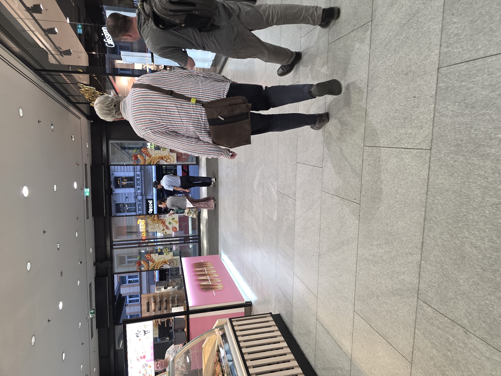The image, taken from a slightly elevated perspective, captures an indoor commercial area, likely a modern shopping mall or food court. The floor is made of large, light gray tiles, and the ceiling features numerous recessed circular lights.
In the mid-ground, two individuals are walking away from the camera. The person closer to the camera has gray hair, wears a red and white striped long-sleeved shirt, dark pants, and carries a brown messenger bag. Behind them is another person with short dark hair, wearing a gray top and gray pants, with a dark backpack.
To the left, a series of glass storefronts line the passage, reflecting portions of the building's interior and the street outside. Inside these storefronts, other people are visible, including one person in a white shirt and dark pants, and another woman in a pink top. Food displays featuring items like waffles or crepes are also visible through the glass. A bright pink display counter with several round, bread-like items on skewers is prominent on the right side of the passage. Another display case with various baked goods is seen in the foreground to the right, with a person's head visible behind it.
Visible text includes a partial sign that reads "Cassius" and another that appears to be "Oishi" (with "SUSHIS" visible in reflection), suggesting food establishments. Green exit signs are also present on the ceiling. The overall scene depicts a well-lit, busy public space during what appears to be daytime.