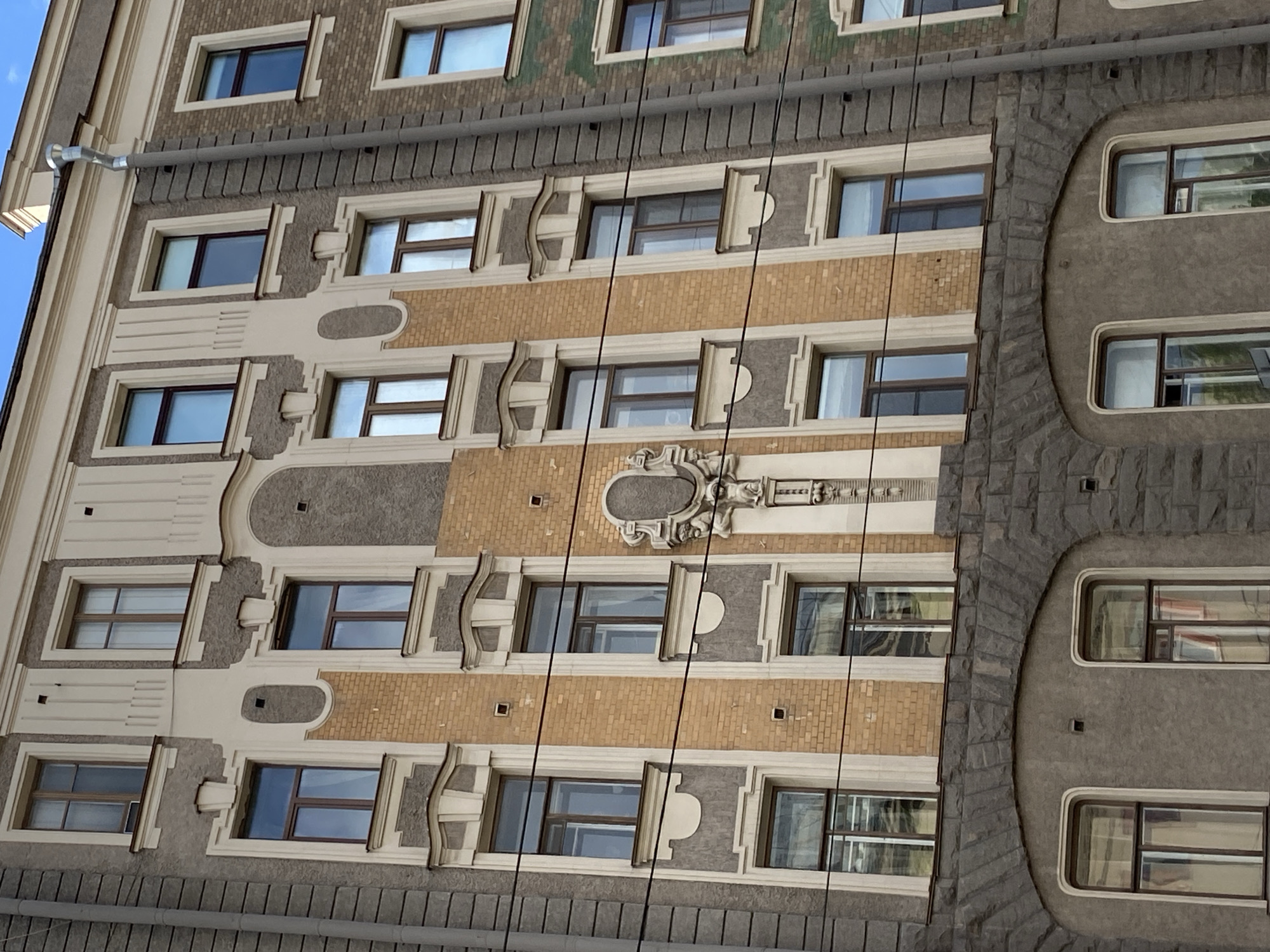 This is a close-up, low-angle shot of a building facade in Saint Petersburg, Russia. The building has a mix of architectural styles, including sections with light-colored brick, decorative stone elements, and varying window designs. The facade is divided into vertical sections with different textures and colors. A gutter runs along the top of the frame. There are black cables running from the top to the bottom of the building. The sky is visible in the upper left corner.