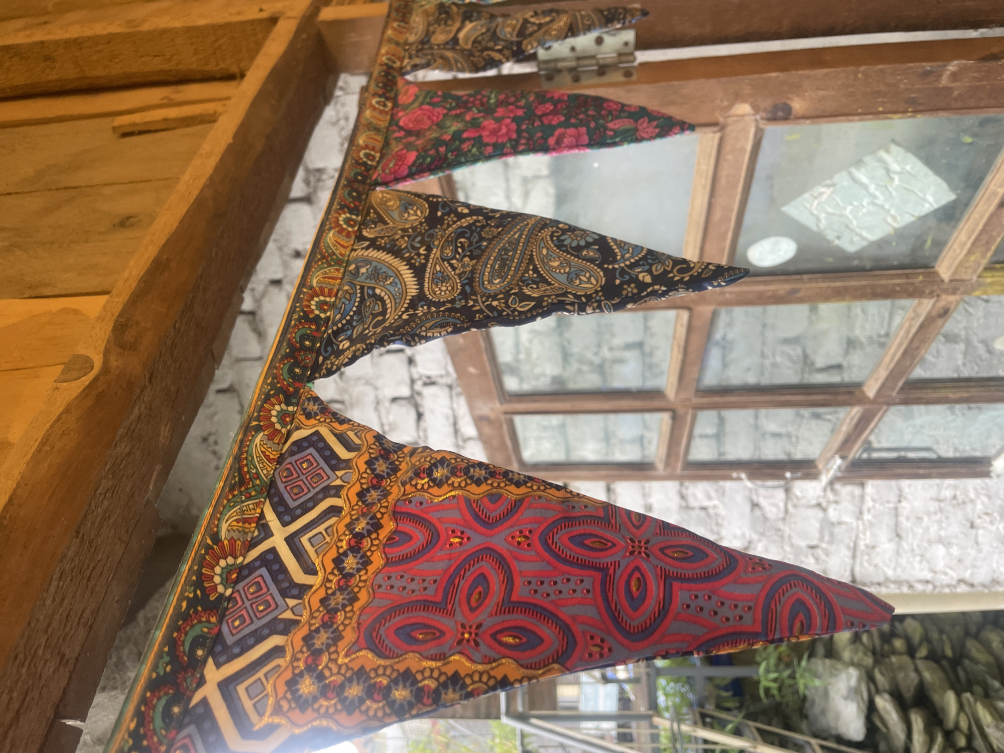A close-up, eye-level, outdoor shot shows a string of colorful fabric pennants hanging from a wooden beam in Bhagsu, India. The pennants are triangular, with intricate patterns and gold accents. The first pennant from the left is predominantly red and gray, with an abstract, geometric design. The second is navy blue with a paisley pattern in shades of blue and tan. The third features a floral pattern of pink roses on a dark background. The fourth pennant is partially visible and appears to have a paisley pattern as well.
Behind the pennants, a rough-textured white brick wall is visible. To the right, a wooden framed window with multiple panes is present, and its glass reflects some light. To the left of the pennants, a metal railing and some greenery can be seen, suggesting an outdoor setting. The overall lighting suggests it is daytime.