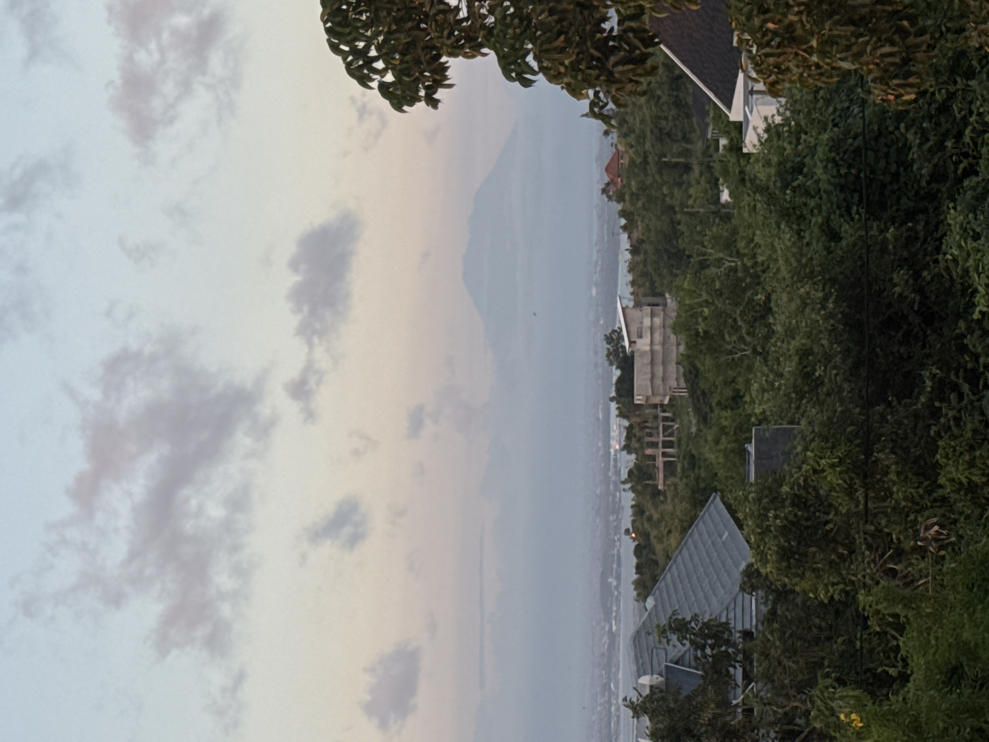 The image shows a view from Ungasan, Indonesia. The sky is filled with a mix of light blue and gray clouds. In the distance, a mountain silhouette is visible. The foreground includes lush green trees and rooftops of buildings. The image is oriented vertically.