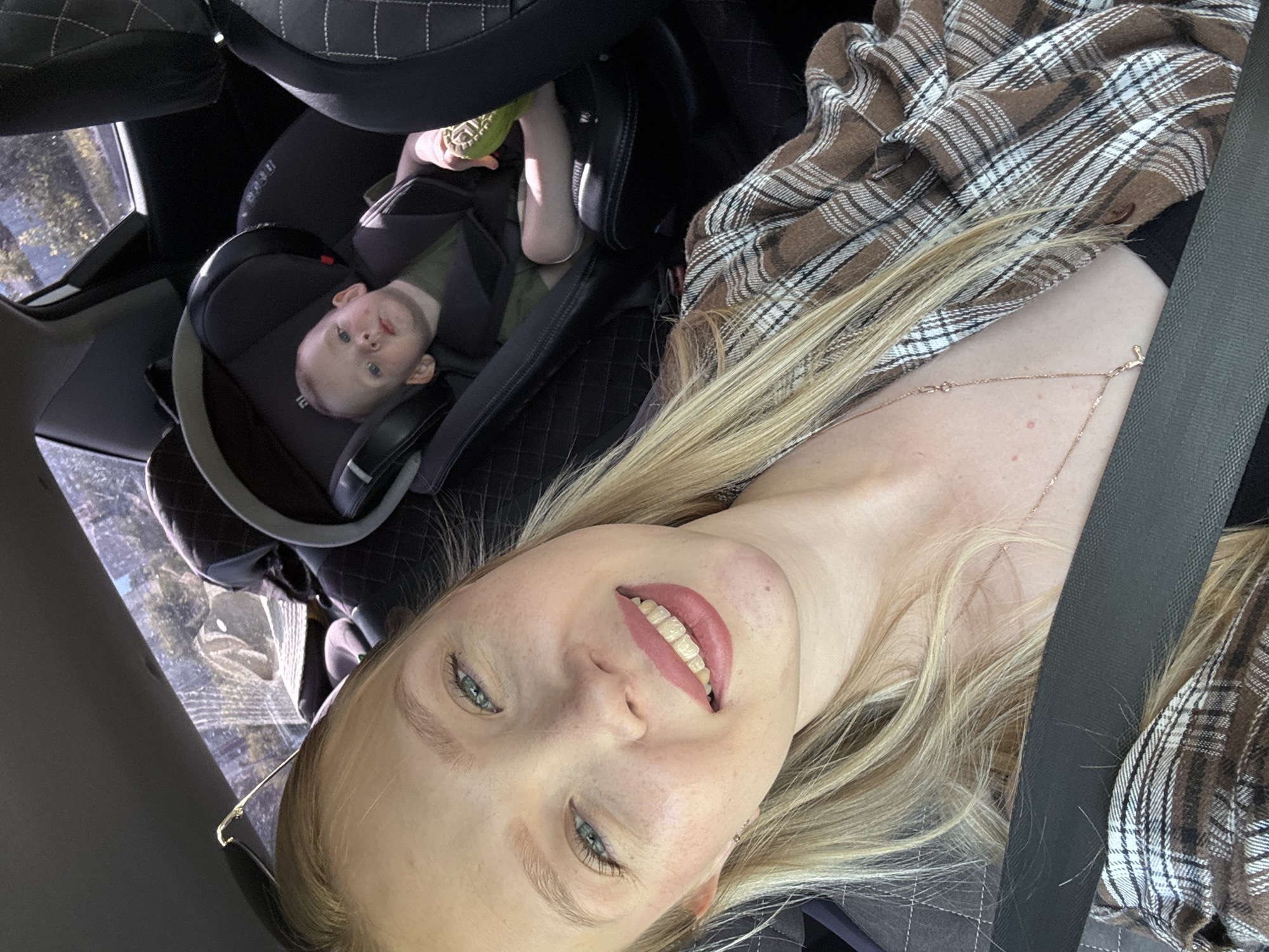 The image shows a woman and a baby in a car. The woman is in the foreground, taking a selfie. She has blonde hair and is wearing a brown and white plaid shirt. Her eyes are blue and she has on lipstick. A thin gold necklace is visible around her neck. The baby is in a car seat behind her, to the left. The baby has fair skin and is looking towards the camera. The baby is wearing a green shirt and is holding a toy. The car interior is visible, with black seats and a seatbelt. Sunlight streams in through the window, casting shadows and highlighting the subjects. The scene appears to be daytime.