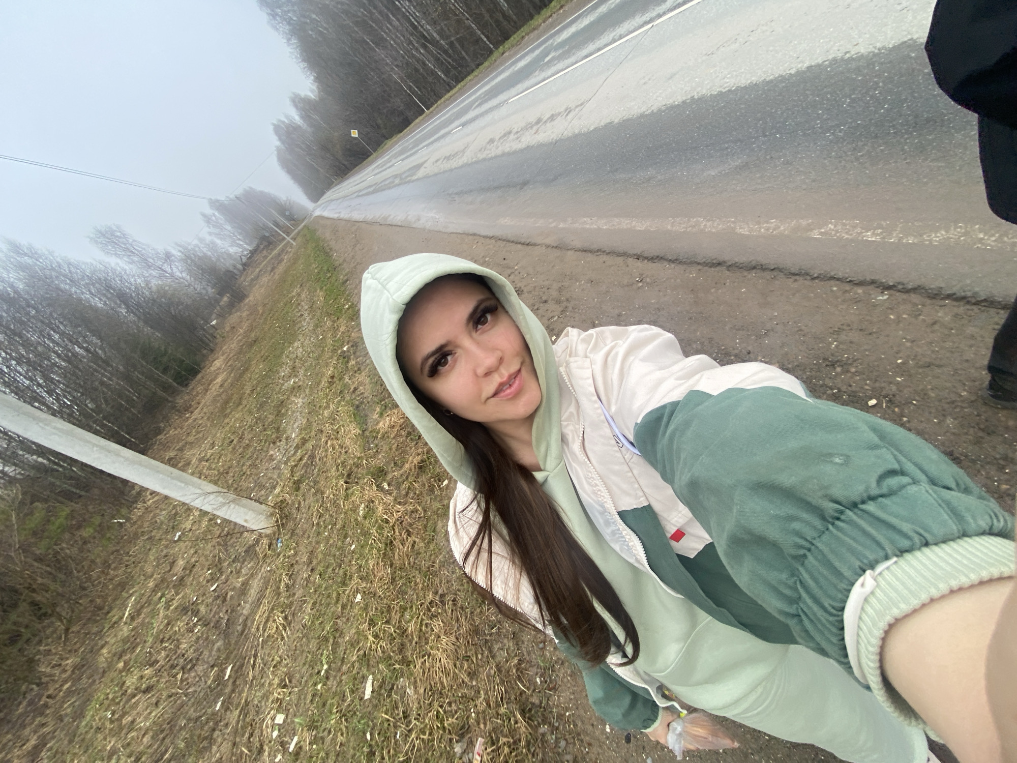The image shows a woman in Zhigalki, Russia, standing on the roadside, possibly taking a selfie. She is wearing a light green hooded sweatshirt and a cream-colored jacket with green sleeves. The background features a wet asphalt road, trees in the distance obscured by fog or mist, and a grassy roadside with utility poles.