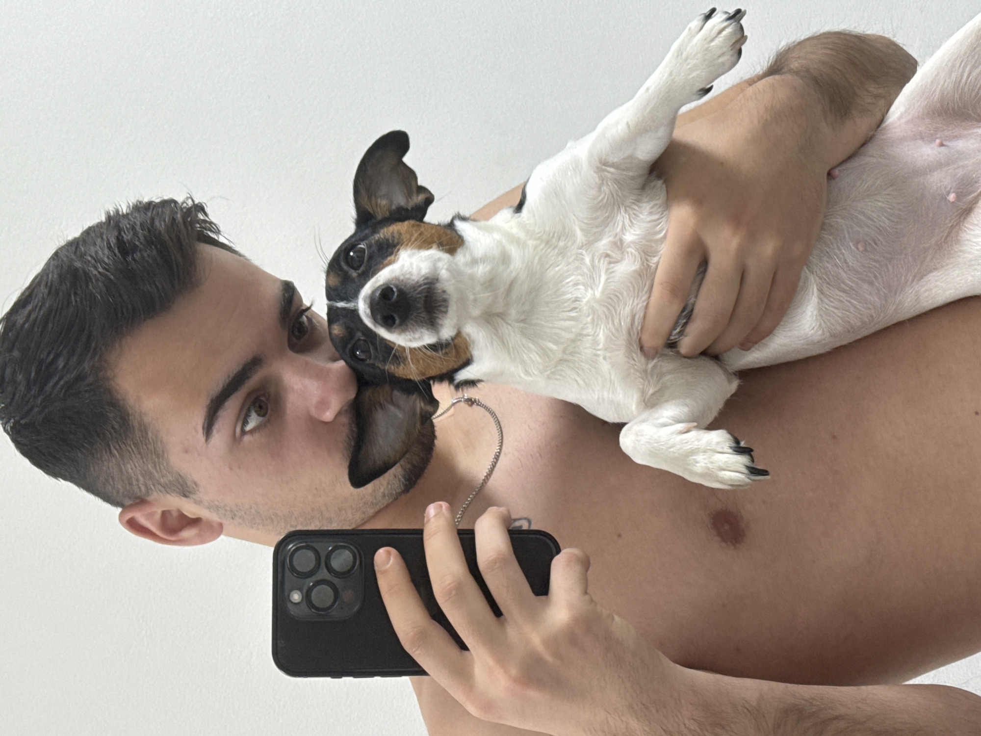 This indoor image, taken in Sofia, Bulgaria, features a young man and a small dog in a selfie-like composition. The man, shirtless with dark hair, light brown eyes, and stubble, is seen from the chest up. He holds a black smartphone, likely an iPhone, in his left hand, angling it towards himself and the dog. A thin silver chain necklace is visible around his neck. His right arm and hand are supporting a small, short-haired dog, possibly a Jack Russell Terrier, which is held upside down against his body. The dog has predominantly white fur with distinct black and brown patches on its head and back, and its pink belly is visible, showing small nipples. Both the man and the dog are looking towards the camera; the man has a neutral to slightly thoughtful expression, while the dog appears alert and curious. The background is a plain white wall, suggesting a simple indoor setting. The lighting is bright and even, indicating a well-lit environment. No visible text is present.