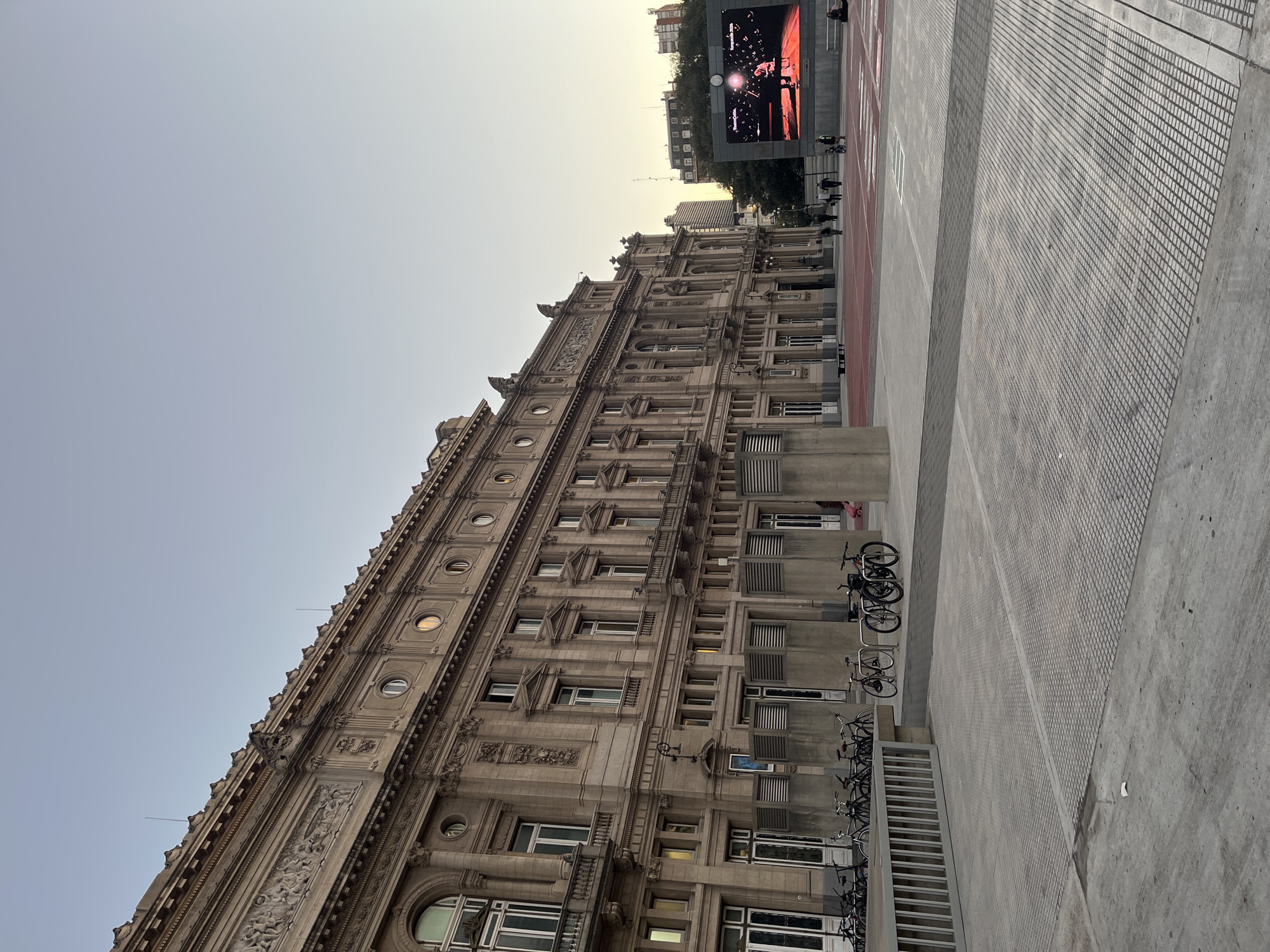 The image captures a grand, ornate building in a city plaza, likely in Buenos Aires, Argentina, given the context. The architecture suggests a historical structure, possibly from the early 20th century, characterized by multiple stories, numerous windows (some round), intricate carvings, pilasters, and cornices. The building's facade is a light, earthy tone, hinting at stone or concrete construction.
Adjacent to the building is a modern urban plaza featuring various ground textures, including smooth concrete, patterned grates, and a reddish track-like pathway. Several rows of bicycles are neatly parked in racks along the base of the building and the plaza, indicating a public bike-sharing or parking system.
In the background, a large LED screen displays what appears to be a performance or event, with a figure on stage and text that partially reads "ESPERANDO" and "GREGORIO". The screen adds a contemporary element to the historic setting. A few indistinct figures of people can be seen walking or standing near the screen and in the distance.
The time of day appears to be late afternoon or early evening, with a clear, light blue sky transitioning to a warmer, lighter hue near the horizon, suggesting the sun is low. The overall scene is calm, depicting an urban environment where historical architecture meets modern city amenities.