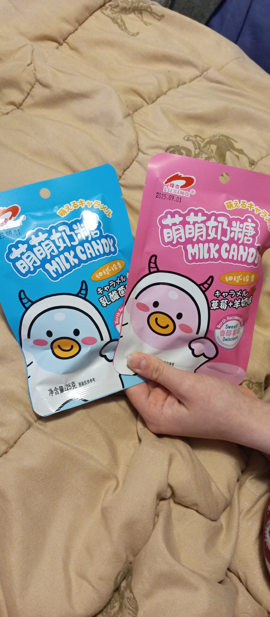 Two bags of "Mengmeng Milk Candy" are shown in a person's hand. One bag is blue, and the other is pink. The bags feature a cartoon cow-like character. The text on the bags includes information about caramel, lactic acid bacteria, and strawberry with goat's milk flavor. The background is a patterned beige blanket. The expiration date is listed as 2025.09.01.