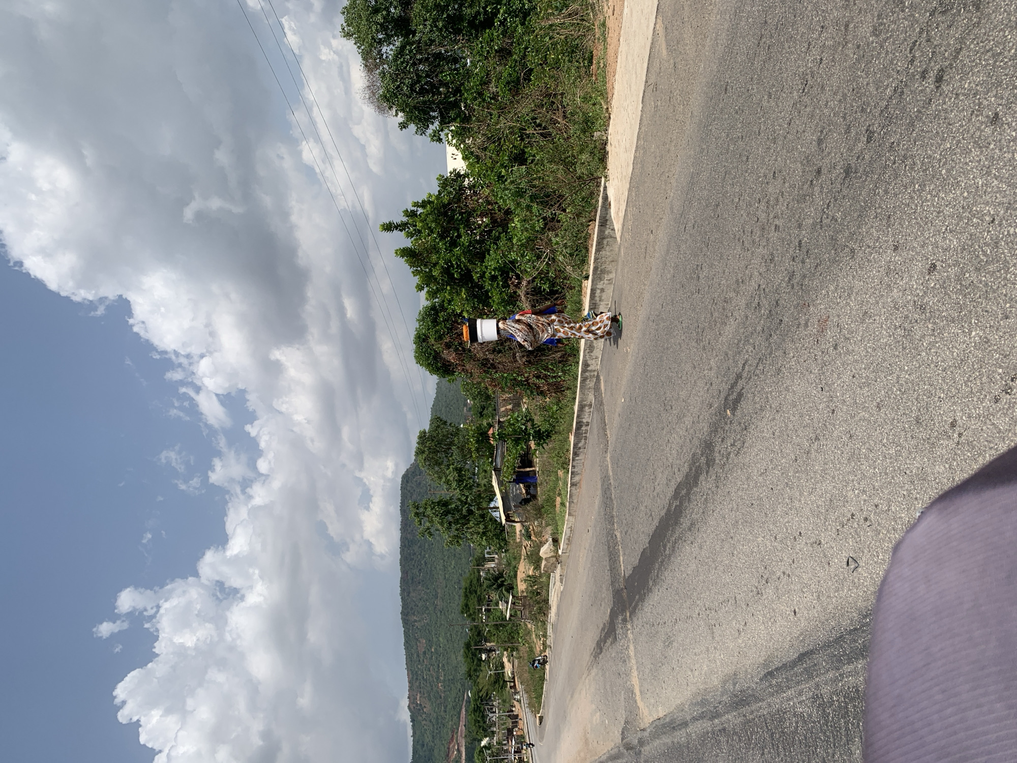 A road runs through a rural part of Lokoja, Nigeria. An adult woman wearing a patterned dress and carrying a white object on her head walks along the edge of the road. Trees line the side of the road. A hillside rises up in the background. The sky is cloudy.