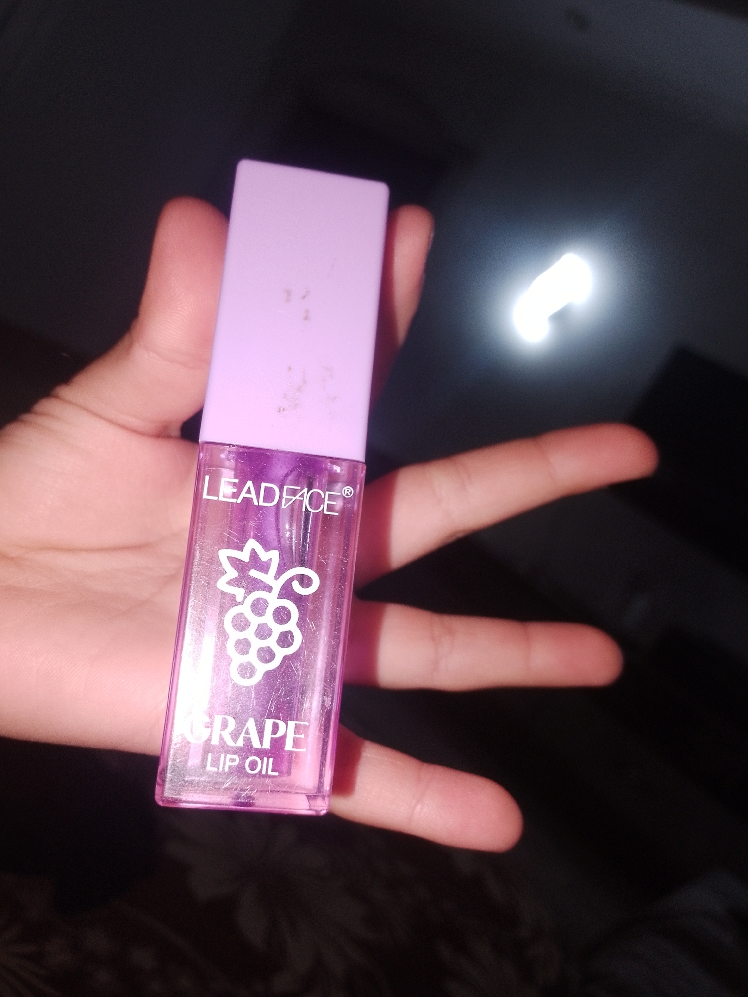 A close-up, dimly lit, slightly blurry, horizontal shot shows a hand holding a small, clear, purple-tinted lip oil tube. The tube has a lavender-colored cap and features a white graphic of grapes and text that reads "LEADFACE" and "GRAPE LIP OIL." The background is dark and out of focus, with a bright, blown-out light source visible in the upper right quadrant, possibly a camera flash or a light fixture. The image appears to be a product photograph taken indoors, with no discernible elements indicating a specific location in the city of Келдемұрат, Kazakhstan.