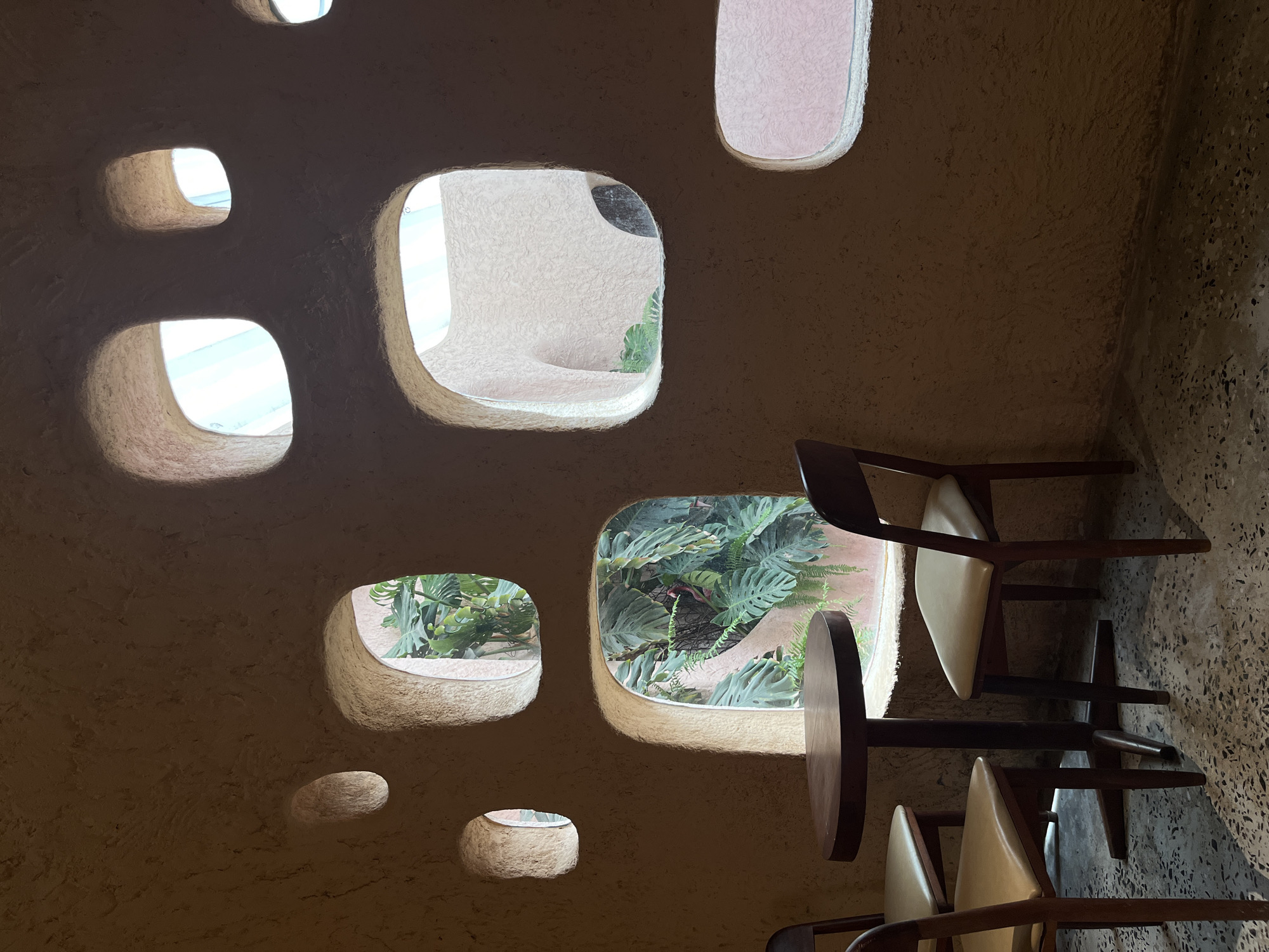 This interior shot depicts an abstract, tan-colored wall punctuated with irregularly shaped, rounded-edge openings revealing exterior views of plants and sky. The wall's texture is rough and uneven, reminiscent of stucco or clay. Through the openings, there's a view of greenery and a hint of light. The right side of the frame shows a section of terrazzo flooring with rows of brown and white chairs. The scene, though artistic, lacks human presence or specific activity, highlighting the architectural features and natural elements. The location, Unknown, Unknown, suggests an unidentified space or city.