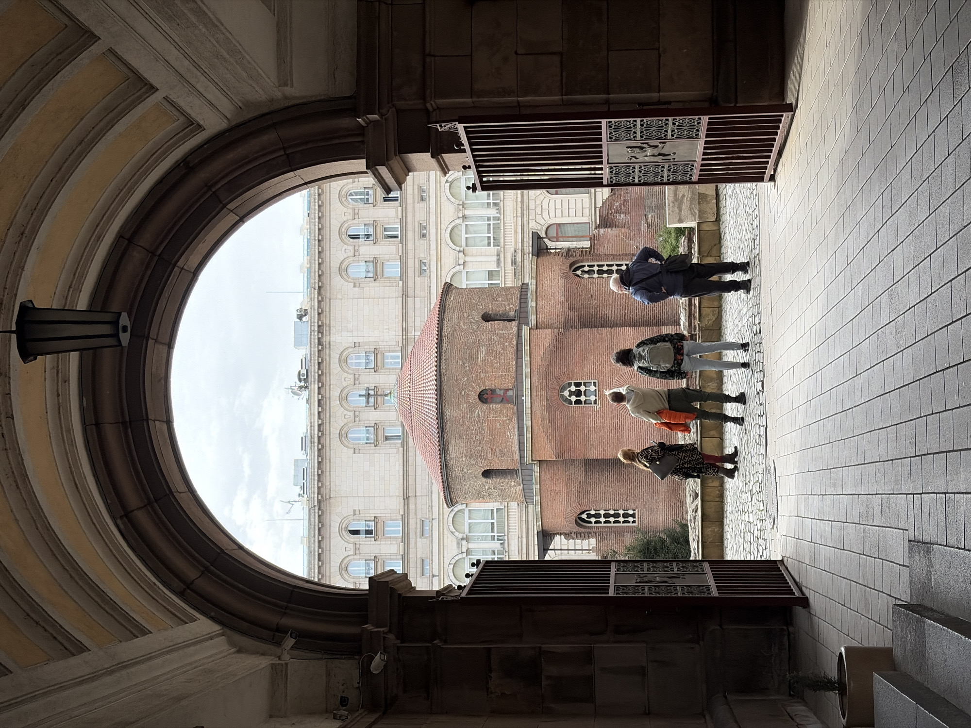 The image presents an outdoor urban scene viewed through a grand, ornate stone archway in Sofia, Bulgaria. The arch, which frames the left and top portions of the foreground, is richly detailed with architectural mouldings and cornices. A dark metal lantern is affixed to the left inner side of the arch.
Beyond the archway, four individuals are visible in the mid-ground, standing on paved ground. From left to right, a person in a light grey jacket and green pants carrying a backpack stands, followed by another in a light top and orange accessory, and a person in a patterned dress or coat. A man in a dark blue jacket and pants with light hair stands slightly apart to the right. They appear to be casually observing their surroundings or waiting, with their gazes directed towards the buildings or down.
The background features two prominent buildings. On the left, a large, light-colored edifice with numerous arched windows spans across multiple floors, reflecting a classic architectural style. To the right, a distinctive brick building with a rounded section and a reddish-brown tiled roof captures attention. This building also showcases varied window designs, including arched and rectangular ones with grid patterns.
The ground immediately outside the archway is paved with rectangular light-colored stones on the right, transitioning to cobblestones or a different pattern closer to the brick building. A decorative metal gate or railing section, featuring an embossed emblem possibly depicting a mythical creature, is attached to the right side of the archway. A small security camera is mounted near the top right of the arch. The sky visible through the arch is overcast, indicating a daytime scene with soft, diffused lighting. No discernible text is visible in the image.