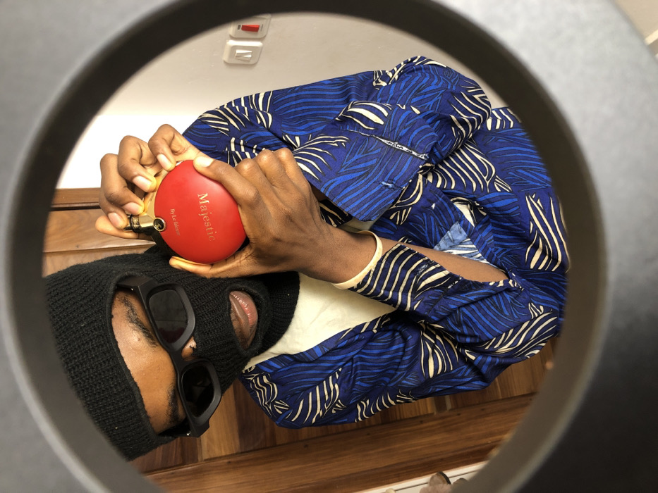 An individual is depicted indoors in Tema, Ghana, framed by a circular aperture. The subject wears a black knitted balaclava, black rectangular sunglasses, and a blue and white leaf-patterned long-sleeved shirt over a light undershirt. Both hands hold a red, circular object with gold-colored metal accents, positioned at mid-torso. The object bears the labels 'Majestic' and 'By Le Chameau'. The individual's mouth is slightly open, and they gaze directly forward. The immediate background comprises a light brown wooden surface on the left and a plain light-colored wall on the right, featuring one red and one white electrical switch or outlet.