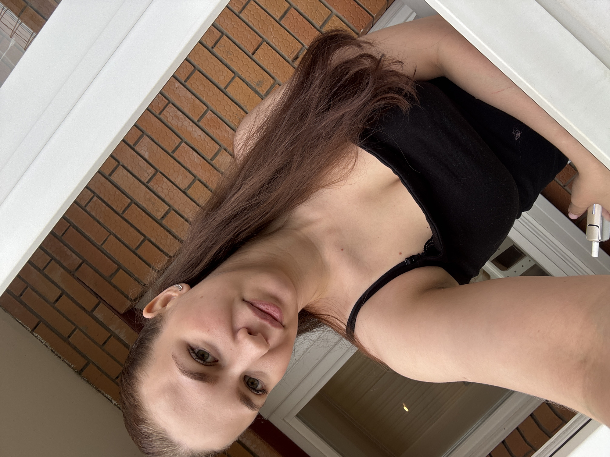 The image is a low-angle selfie of a young woman, likely in her 20s or 30s, taken from what appears to be a window or balcony in Chaikovsky, Russia. The scene frames her within a white structural element, possibly a window or door frame, against a reddish-brown brick wall.
The woman has long, dark brown hair flowing over her left shoulder. Her face is turned slightly towards the camera, with light-colored (green or hazel) eyes and a calm, subtle smile. She is wearing a black top, possibly a camisole or tank top, with a thin strap visible on her right shoulder and a hint of lace near the neckline. A small, silver-toned hoop earring is visible in her left ear. Minor red marks or scratches are noticeable on her right forearm, and her nails are painted a light pink or nude color.
In her right hand, she holds a light-colored object that appears to be a vape pen or electronic cigarette. The background consists of a textured brick wall with brownish-red bricks, indicating an exterior building facade. A blurry reflection in a glass pane suggests an outdoor daytime setting. There is no visible text in the image. The overall impression is one of a casual, self-taken portrait.