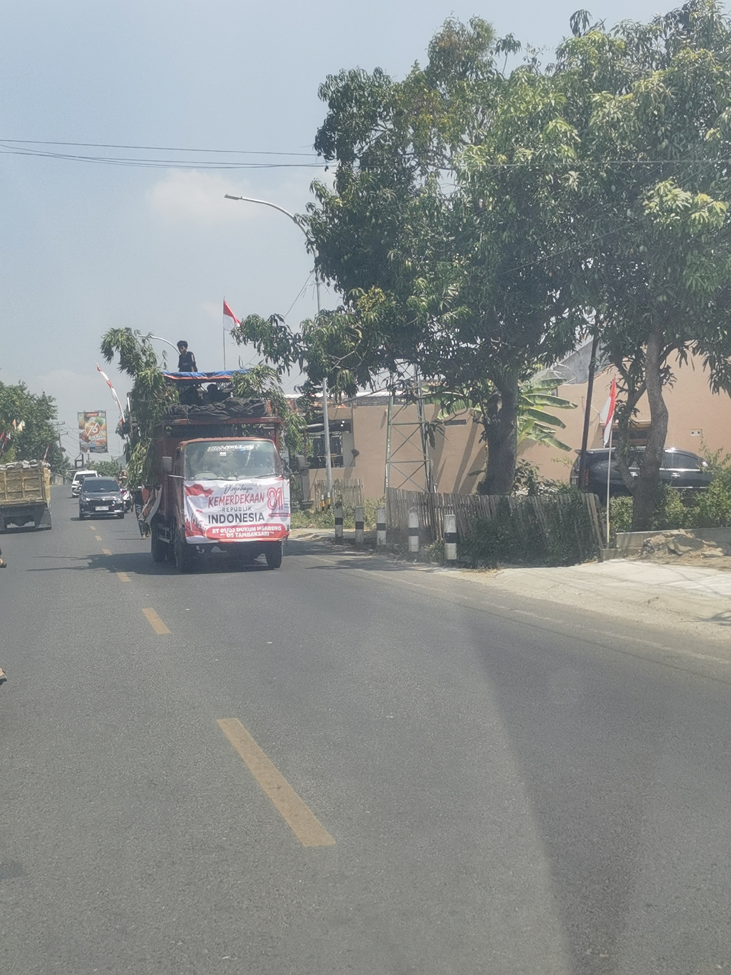 A truck decorated with greenery and a banner reading "Dirgahayu KEMERDEKAAN REPUBLIK INDONESIA 81" is driving down a road in Tambaksari, Indonesia. The truck has a person standing on top of it. The banner also includes the text "RT 01/02 DUKUH NGARANG - DS TAMBAKSARI". There are other vehicles on the road, including cars and another truck. The environment is a street with trees and buildings on the side. The sky is clear and blue.
