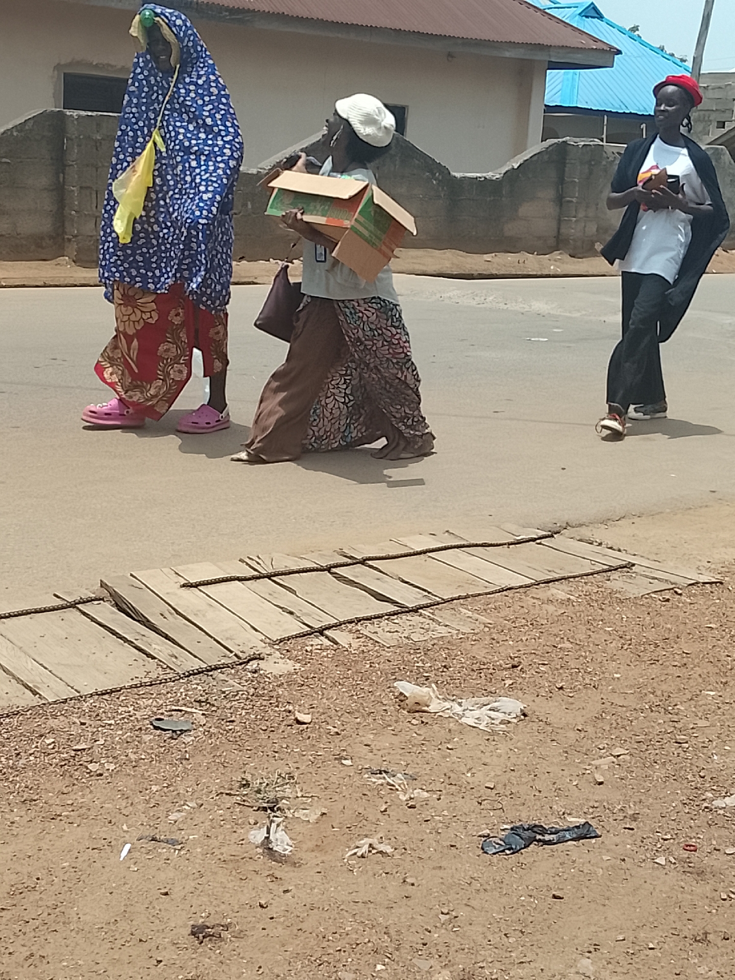 Three individuals walk along a paved road in Akwanga, Nigeria. The first person is wearing a blue and white patterned hooded garment over a floral print skirt and pink Crocs. A yellow bag hangs from their shoulder. The second person carries several cardboard boxes and a purse, wearing a white hat and a printed skirt. The third person wears a red hat, a white shirt, black pants, and a black shawl, holding something in their hands. The road has a wooden cover with chains across a ditch. There is trash on the ground. The buildings in the background have metal roofs.