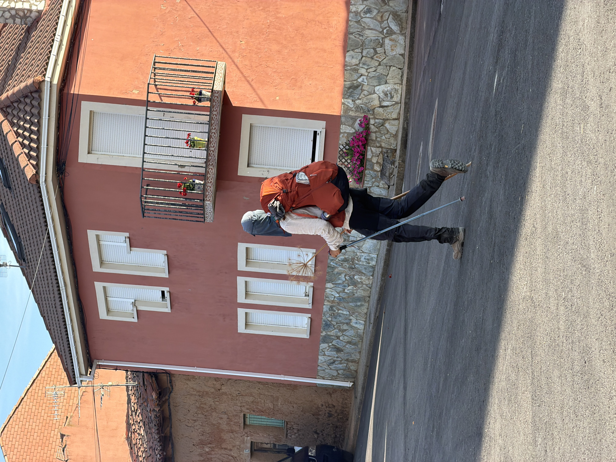 A person with a large orange backpack is walking on a street in Arcahueja, Spain, using trekking poles. The person is wearing a light-colored long-sleeved shirt, dark pants, and hiking boots. They are seen from the side, moving away from the camera. Behind the person is a rustic building with an orange facade and a stone base. The building has several windows with white shutters and a small balcony with potted red flowers. To the left of this building, another building with a brick facade is partially visible. The street is paved and appears to be in a rural or semi-rural setting, with a clear blue sky overhead. The sun is casting shadows on the street. A cat is visible near the stone wall of the orange building.