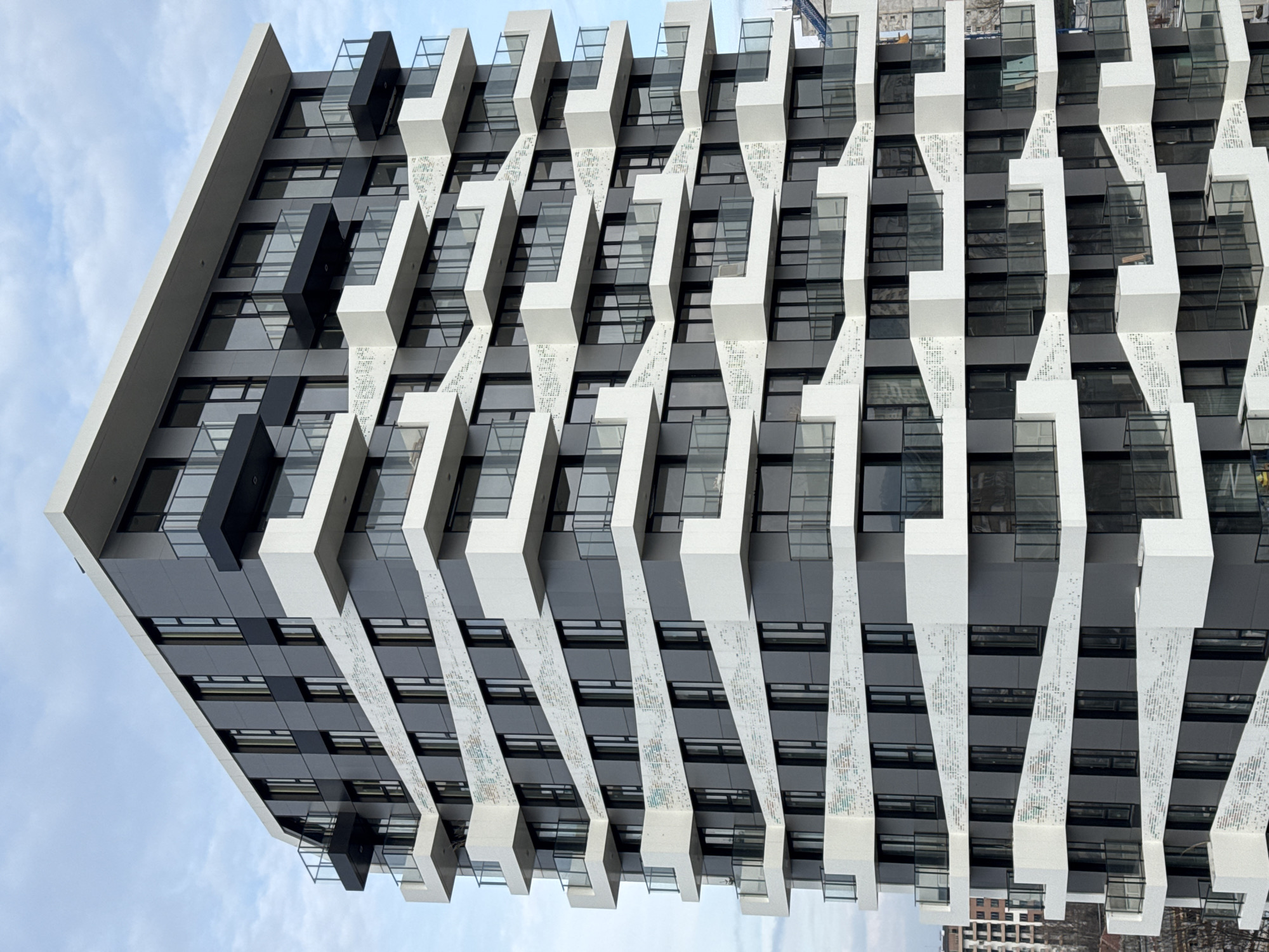 A low-angle, tilted-view shot captures a contemporary architectural structure, presumably a residential or commercial building, located in Sofia, Bulgaria. The building's facade showcases a striking contrast between dark-tinted glass windows set within a grid-like pattern and bold, white, geometric protrusions that extend outward. These white structures have a patterned texture or design element that appears to be a series of small perforations. At intervals, some windows are augmented with black, box-like balconies that jut out from the main structure. The building's left side is distinguished by a smooth, white edge that runs vertically along the length of the building. The sky is visible in the lower left of the image, suggesting that the shot was taken from ground level. In the distance, towards the bottom of the frame, portions of neighboring buildings are visible.