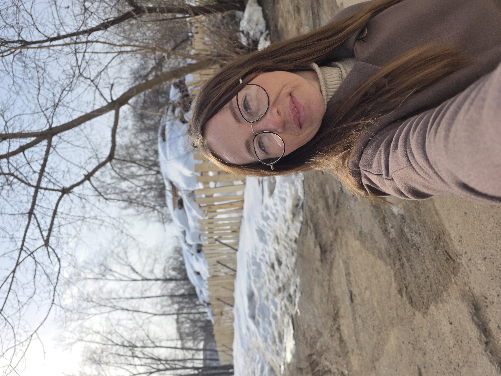 An outdoor selfie taken in Chaikovsky, Russia, captures a female subject positioned in the foreground. The woman has long brown hair, wears round eyeglasses with thin metal frames, a light beige turtleneck, and a brown overcoat. She exhibits a subtle smile and holds the camera device with her extended right arm. The background reveals a dirt path in the immediate foreground, showing vehicle tire tracks and patches of melting snow and mud. Beyond the path, a light-colored wooden picket fence spans horizontally, partially submerged in substantial snowdrifts. Numerous bare deciduous trees are visible behind the snow and fence, under a bright, light blue sky. The overall environment suggests a semi-urban or residential fringe area during late winter or early spring. No other individuals are present.
