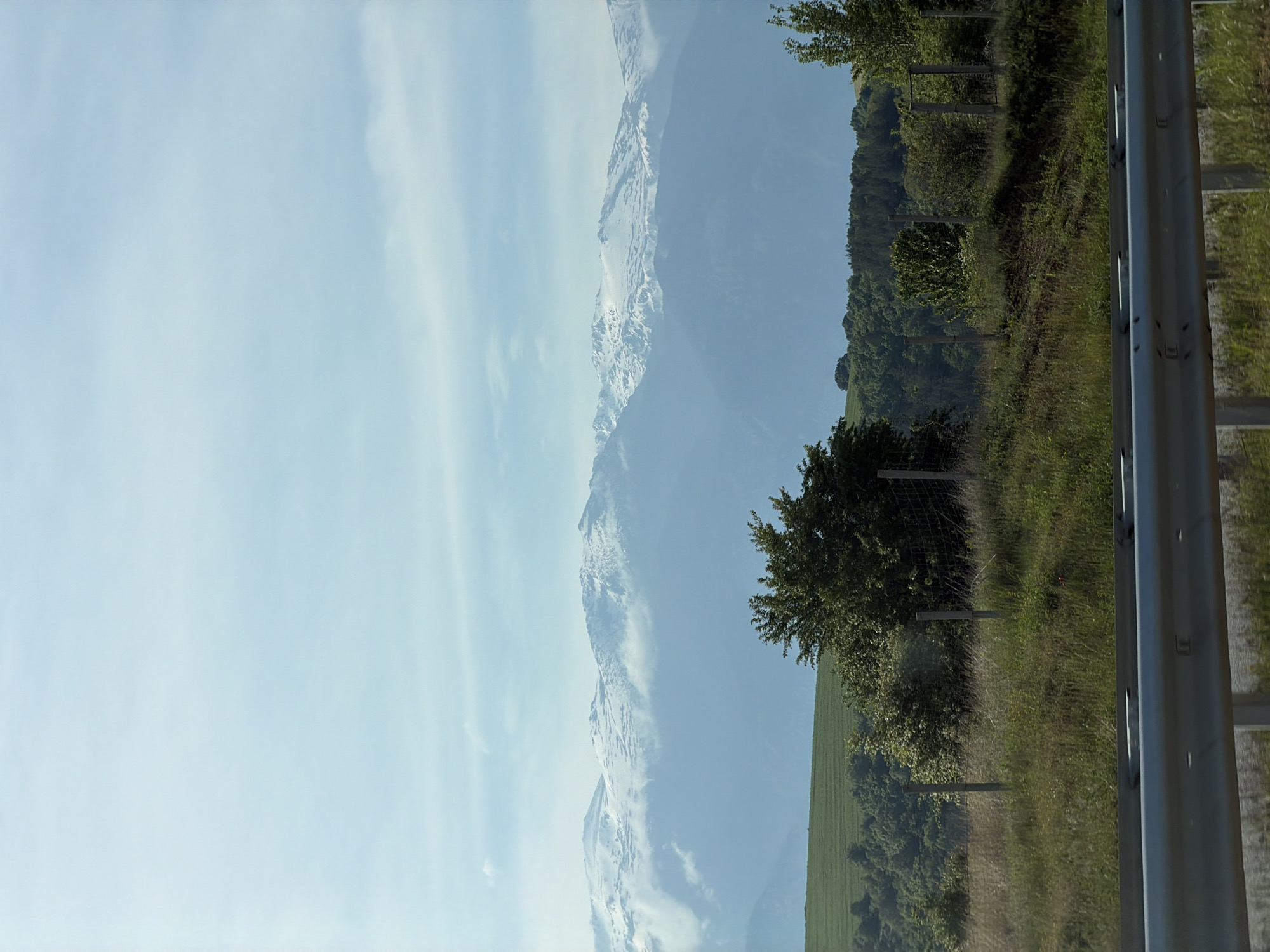 The image presents a landscape possibly observed from a vehicle moving along a road, evidenced by the presence of a metal guardrail on the right side. To the left, a broad expanse of sky occupies the upper portion of the scene, gradually transitioning into a range of mountains veiled by atmospheric haze, some of which retain patches of snow. Adjacent to the right of the mountain range is a terrain exhibiting undulating green vegetation. A line of trees, intermittently spaced with fencing posts, demarcates a boundary within this vegetation, bordering a further area of green land. The general landscape may be observed near Dyakovo, Bulgaria based on metadata.