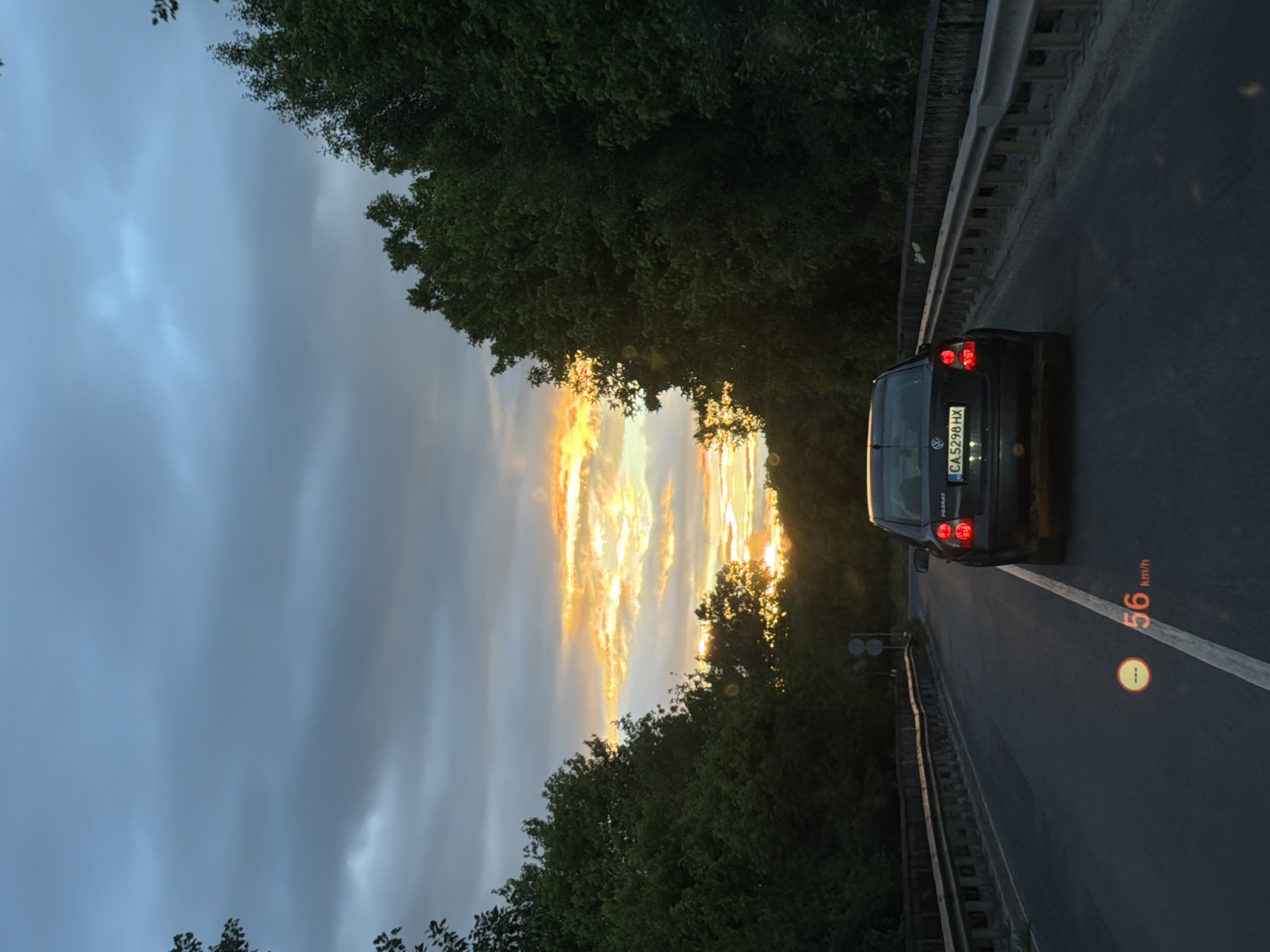 The image depicts a black Volkswagen Passat driving on a road in Katunitsa, Bulgaria. The car's license plate reads "CA 5298 HX." The speedometer indicates a speed of 56 km/h. The road is lined with trees and has a guardrail on the side. The sky is cloudy with a glimpse of the setting sun, casting a golden glow through the trees.
