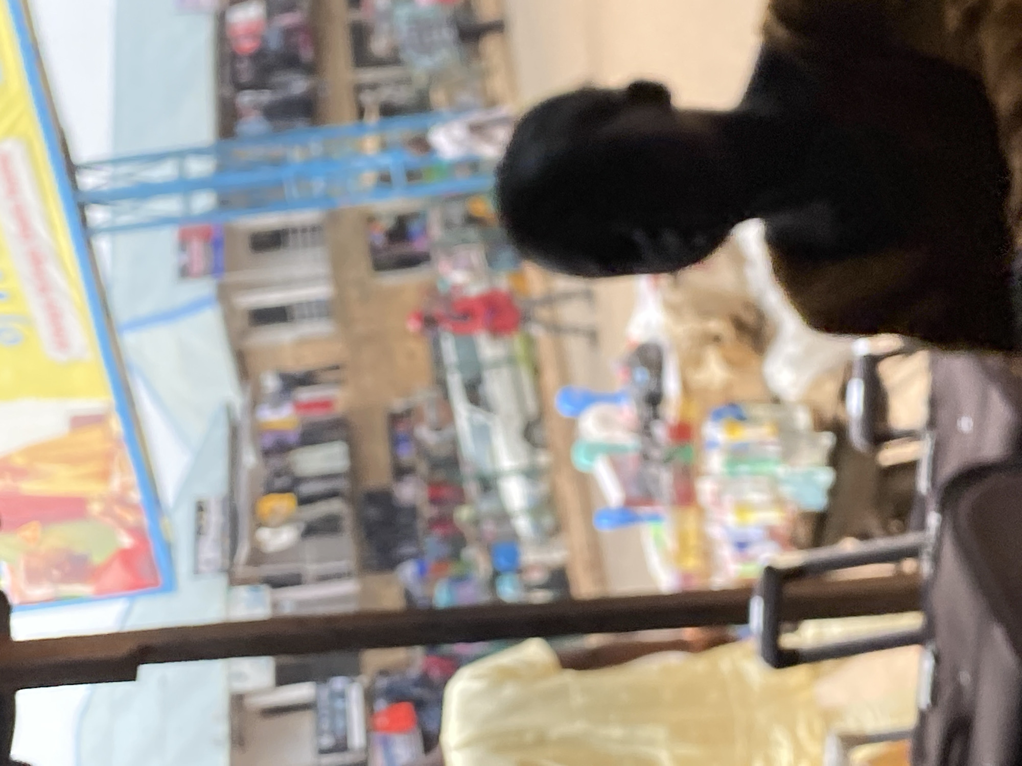 A slightly blurred, medium shot captures an outdoor scene in Bukuru, Nigeria. The foreground is dominated by the upper portion of dark-colored luggage. To the left, a person wearing a bright yellow garment is partially visible. In the mid-ground, a stall displays an assortment of colorful items, possibly drinks in bottles and plastic containers. Behind the stall, several individuals are visible, including one in a red garment walking towards the right, and others further in the background. A white vehicle is parked in the middle distance. A large, bright yellow banner with text and images is affixed to a blue metal structure overhead, suggesting a commercial or public area. The environment appears to be a street or market setting, with buildings and a general sense of activity. The lighting suggests it is daytime.