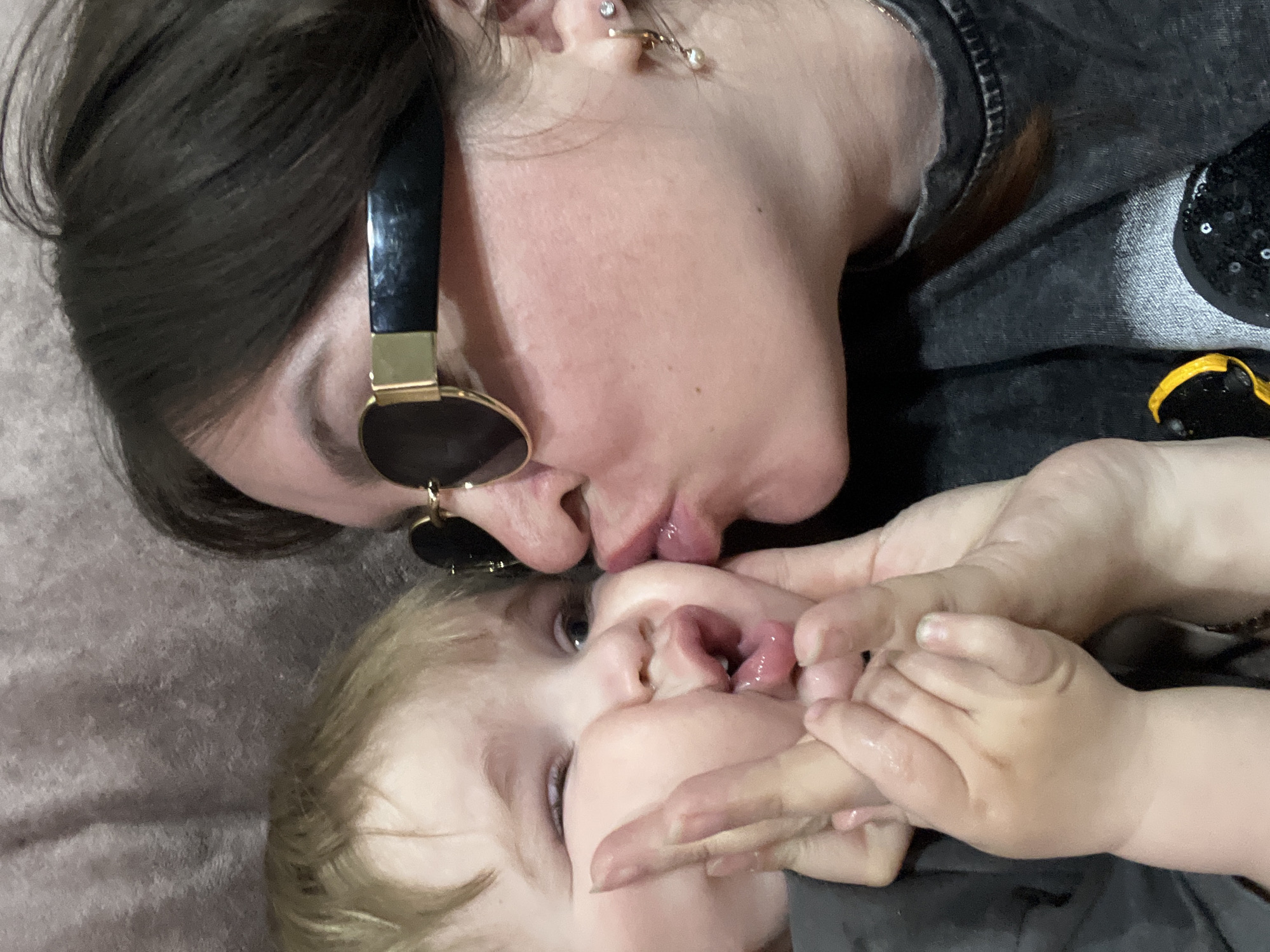 A light-skinned woman with dark hair, wearing black sunglasses and a dark gray shirt, is lying down and kissing a light-skinned toddler with light blonde hair. The toddler is lying on his/her back. The woman's right hand is holding the toddler's hands. The location is not mentioned in the image and cannot be determined.
