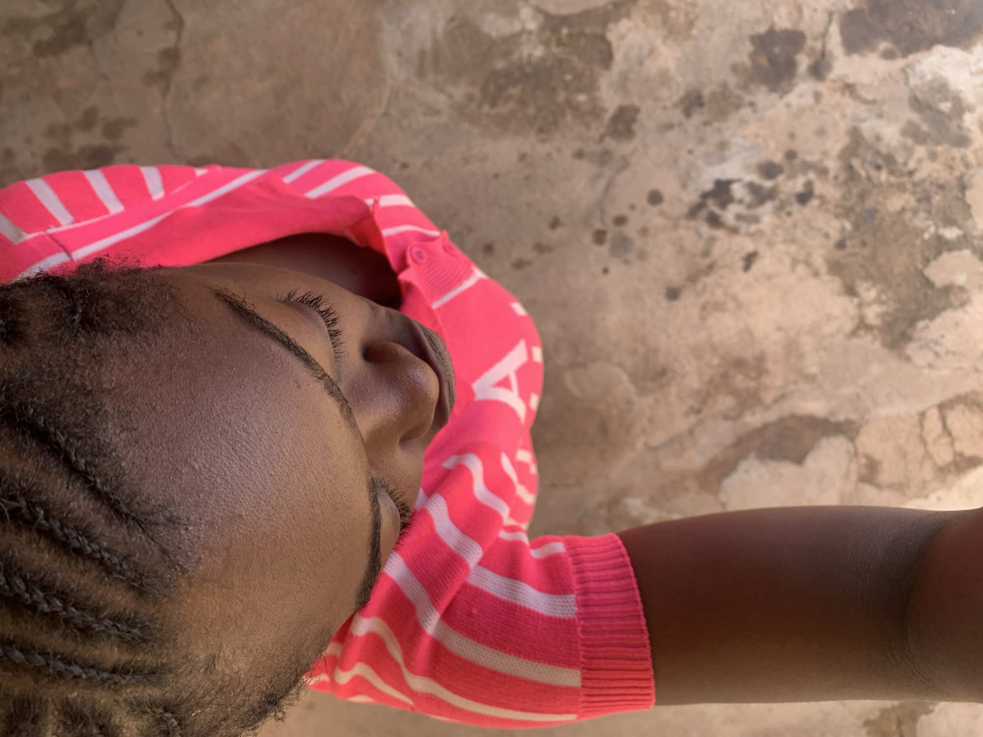An overhead photograph, taken in Rafin Par, Nigeria, depicts a person looking downwards. The subject has dark skin and tightly braided hair on the top of their head. They are wearing a bright pink long-sleeved top featuring multiple white horizontal stripes of varying widths. Part of a white letter "A" is visible on the upper arm/shoulder area of the garment. The person's eyes are mostly closed or cast downwards. The lower part of one arm, clad in the pink and white striped sleeve with a ribbed cuff, extends towards the bottom left of the frame. The background is a textured, light brown or tan ground surface, exhibiting irregular dark stains, minor cracks, and rough patches, consistent with outdoor pavement or packed earth.