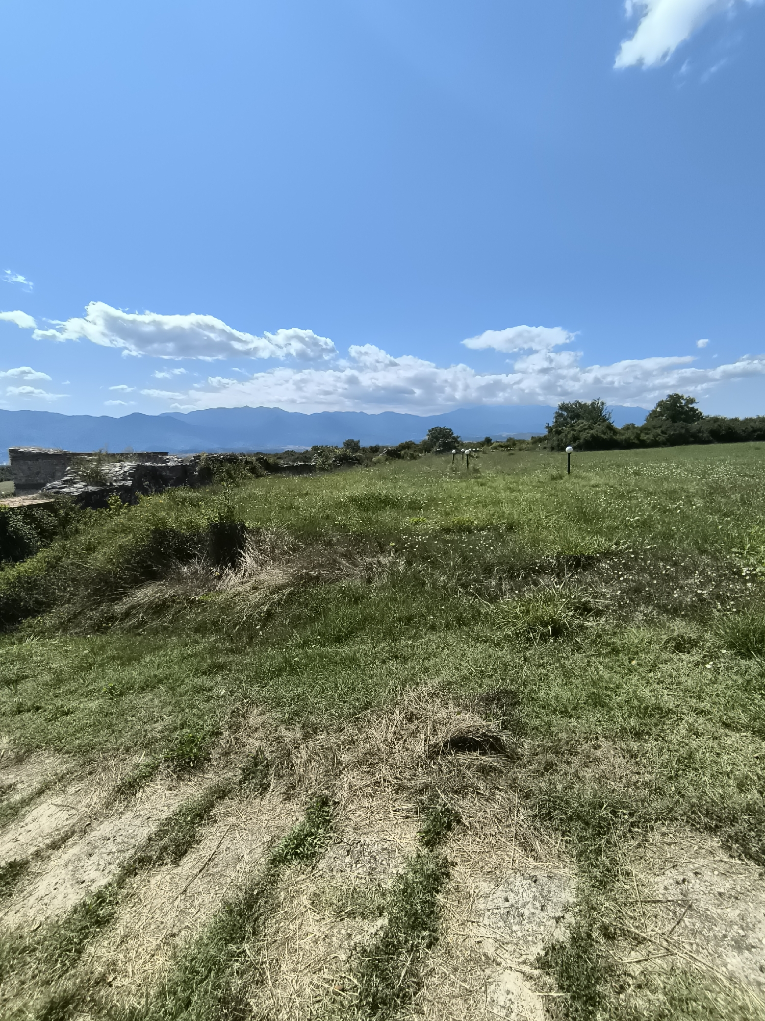 The image depicts a landscape scene with a clear blue sky dotted with white clouds. In the foreground, there is a grassy hill with dry straw and patches of bare earth. The middle ground features a field of green grass with scattered small white flowers. Several white spherical lights on dark poles are visible in the field, forming a line. In the background, there are trees and distant mountains. On the left side of the middle ground, there are ruins of a stone structure. The scene appears to be an outdoor location in Garmen, Bulgaria.