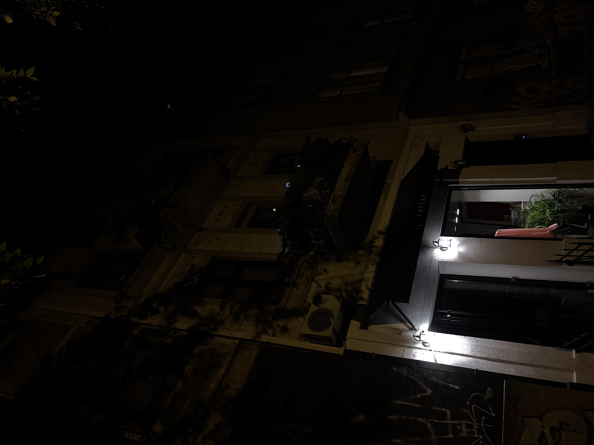 An upward shot shows a building facade in Belgrade, Serbia, at night. The building's lower portion, possibly a ground-level entrance or facade, has graffiti. An air conditioning unit is visible, protruding from the wall. One of the upper levels is brightly lit, revealing plants and a pink item on a balcony. A sign bearing the text "ja tarlač" is visible next to the illuminated balcony. The left portion of the image is obscured by darkness and foliage. The building's architecture features varying window and wall textures.