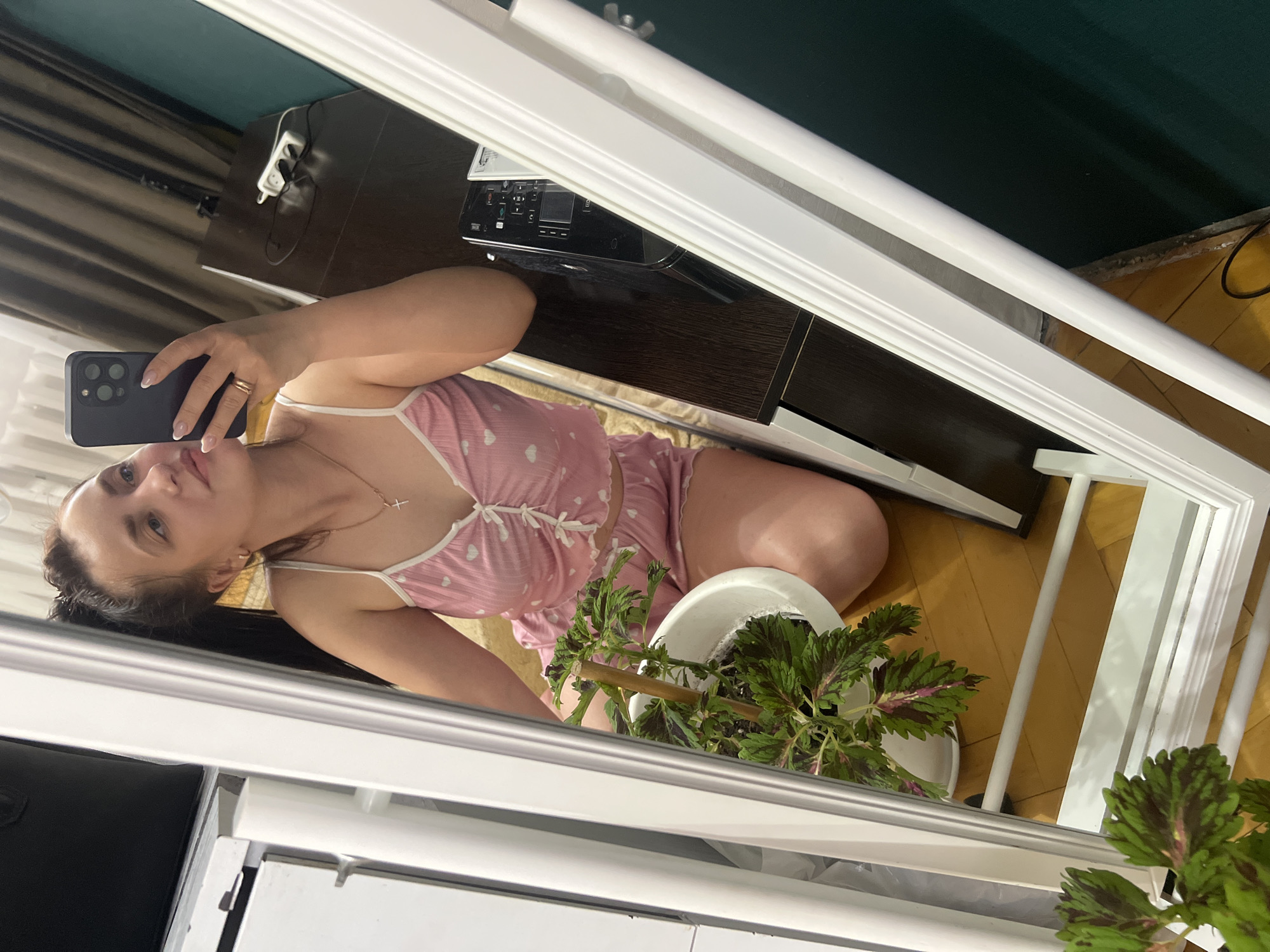 A young woman with her hair in a high ponytail is kneeling on a wooden floor, taking a mirror selfie with her phone. She is wearing pink pajamas with a heart pattern and a gold necklace with a cross pendant. A potted plant with green and purple leaves is to her left. In the background, a dark wooden desk holds a black electronic device and a white extension cord with plugs. The scene appears to be indoors, likely in a bedroom or living area. There is no specific indication of the location being in Марковский, Russia.