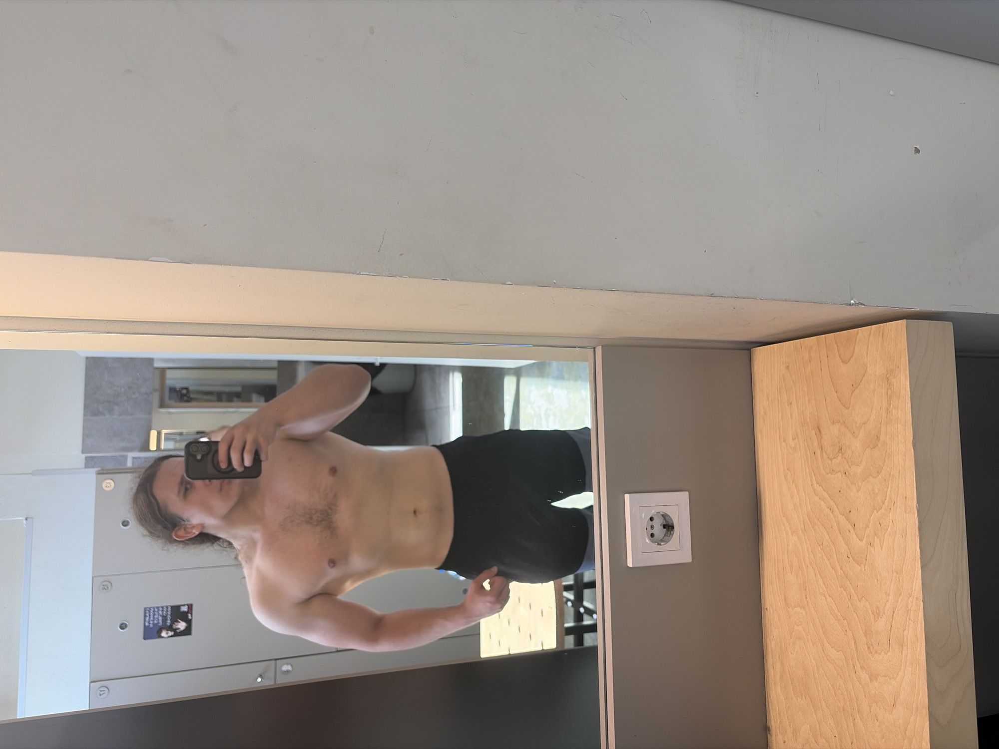 A shirtless man is taking a selfie in front of a mirror. The man is fair-skinned and has long brown hair. He is wearing black shorts. The mirror reflects a locker room or similar space with lockers and what appears to be a bathroom in the background. An electrical outlet is visible on the wall next to the mirror. A wooden panel is situated to the right of the mirror. There is no information in the image that would indicate the location as Plovdiv, Bulgaria.