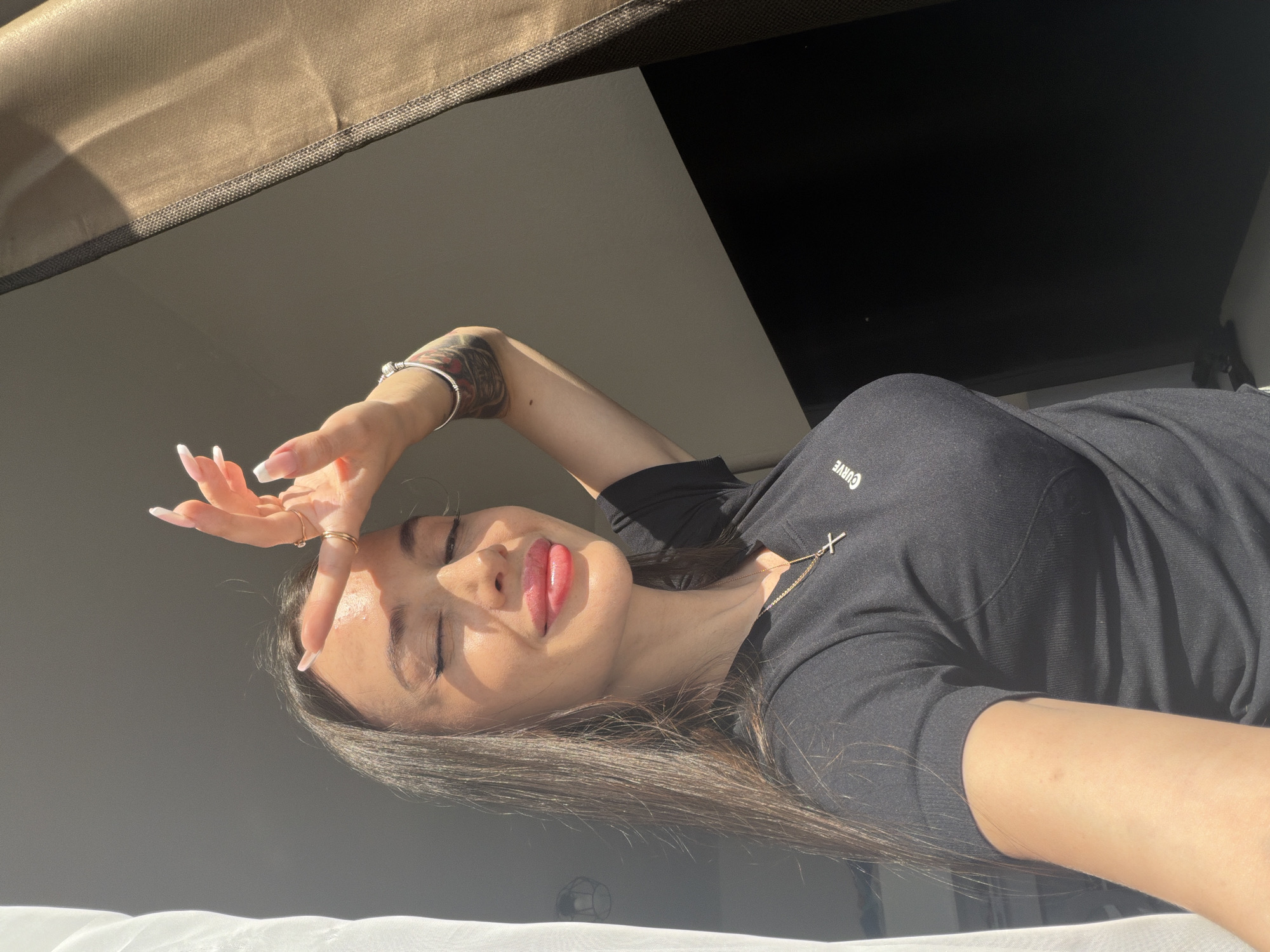 The image displays a young woman in an indoor setting, positioned on her side with her head facing upwards. She has long dark hair and is wearing a black short-sleeved top with the word "CURVE" printed in white on the front, near her left shoulder. A gold chain necklace with a white cross pendant is visible around her neck.
Her left hand is raised, with fingers spread, shielding her eyes from direct, bright sunlight. Her eyes appear closed or squinting due to the light, and her lips are painted with a vibrant red or pink lipstick, forming a slight, closed-mouth smile. She has long nails with white French tips and wears multiple gold rings on her left hand, along with a silver bracelet on her left wrist. A tattoo with intricate patterns is visible on her left forearm.
The setting appears to be a room bathed in bright natural light, casting distinct shadows and highlighting her features. The background consists of a dark, ambiguous area behind her upper body, contrasting with a lighter, possibly white, surface below her, which could be a bed or cushion. The overall atmosphere suggests a relaxed, daytime moment. The provided context indicates the location is Saint Petersburg, Russia.