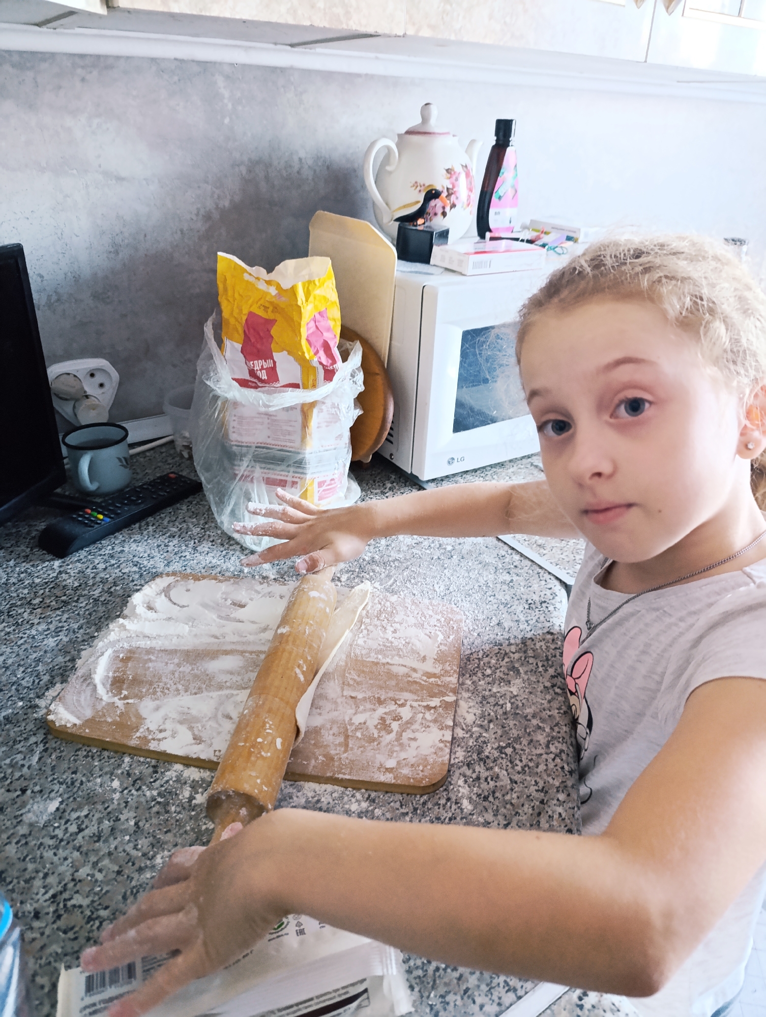 In an indoor kitchen setting, a young Caucasian girl with blonde hair is pictured. She is wearing a grey t-shirt with a cartoon character print and a silver necklace. Her hands are covered in flour as she is in the process of rolling out dough with a wooden rolling pin on a wooden board. The kitchen counter is speckled grey, and a bag of flour is visible in the background, along with a microwave oven, a teapot, and other kitchenware. The overall scene suggests a domestic activity, likely baking.
