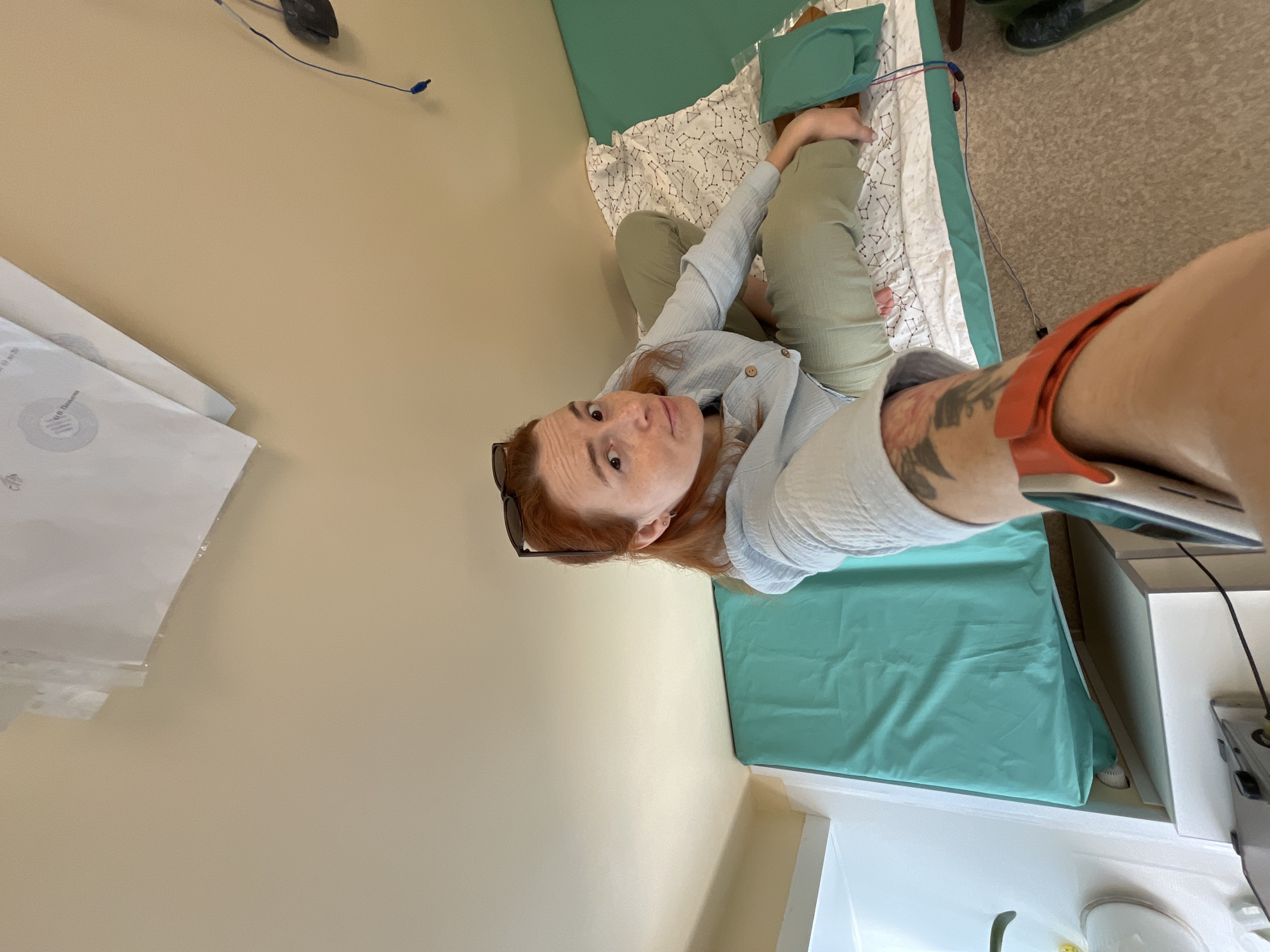A first-person perspective selfie shows a woman with auburn hair, wearing sunglasses on her head, a light blue button-down shirt, and olive green pants. She is sitting on a bed with a turquoise sheet and a white sheet patterned with constellations. The room appears to be a hospital or clinic room with beige walls. To the left, part of a white medical cabinet is visible. A black cord with red and blue wires is seen connected to the woman's leg, and a smartwatch with an orange band is on her wrist. The background shows a wall with some papers attached to it. The image appears to be taken indoors, in what seems to be a medical facility in Chaikovsky, Russia.