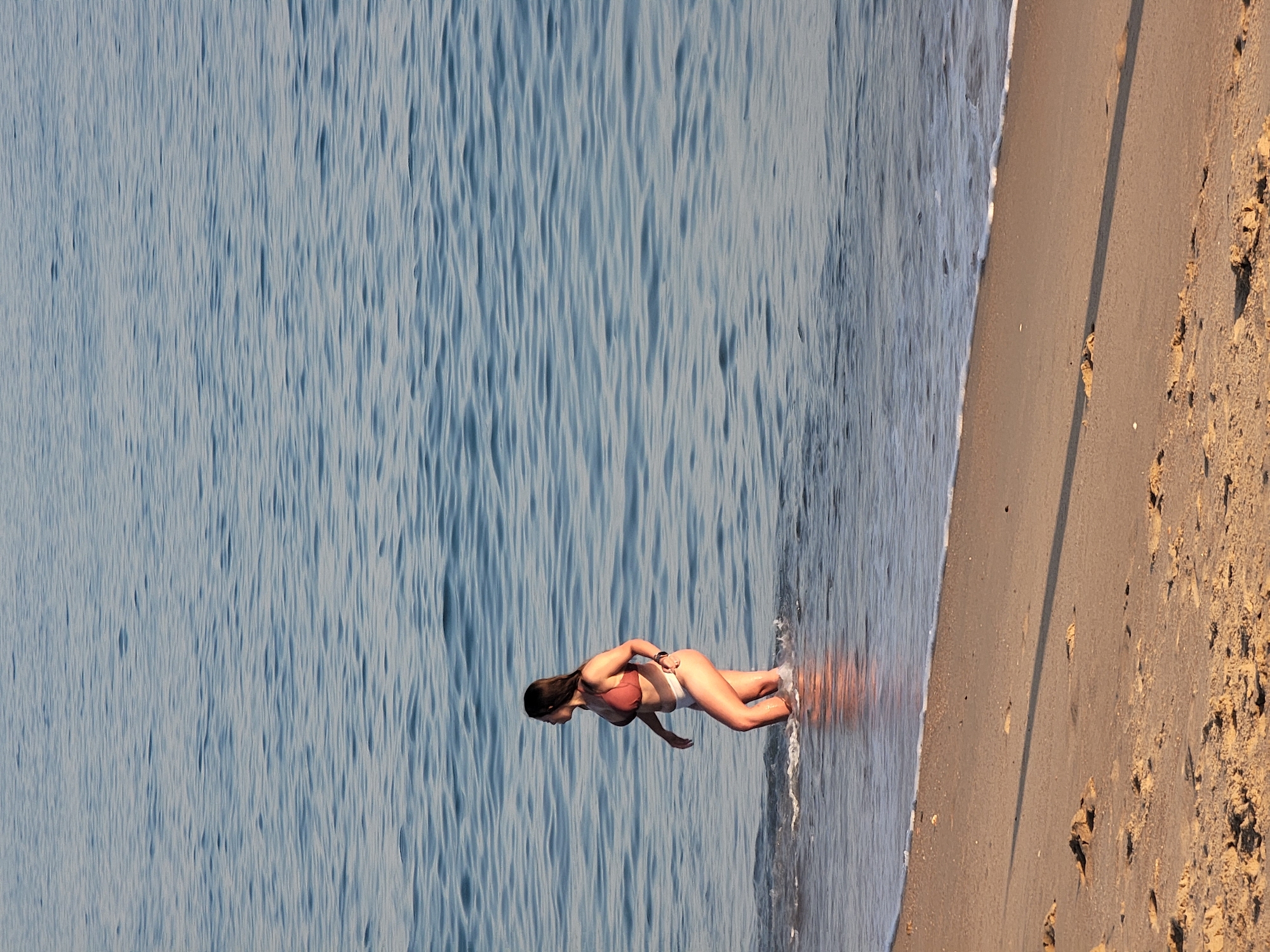 A woman in a bikini is walking in the shallow water at the edge of a sandy beach in Quarteira, Portugal. The water is calm with small ripples, and the sand is light brown with some footprints and debris visible. The woman has long dark hair and is wearing a dark orange bikini top and white bikini bottoms. She is facing left and appears to be looking out at the water. The sun is casting a shadow on the sand, indicating it is either morning or late afternoon.