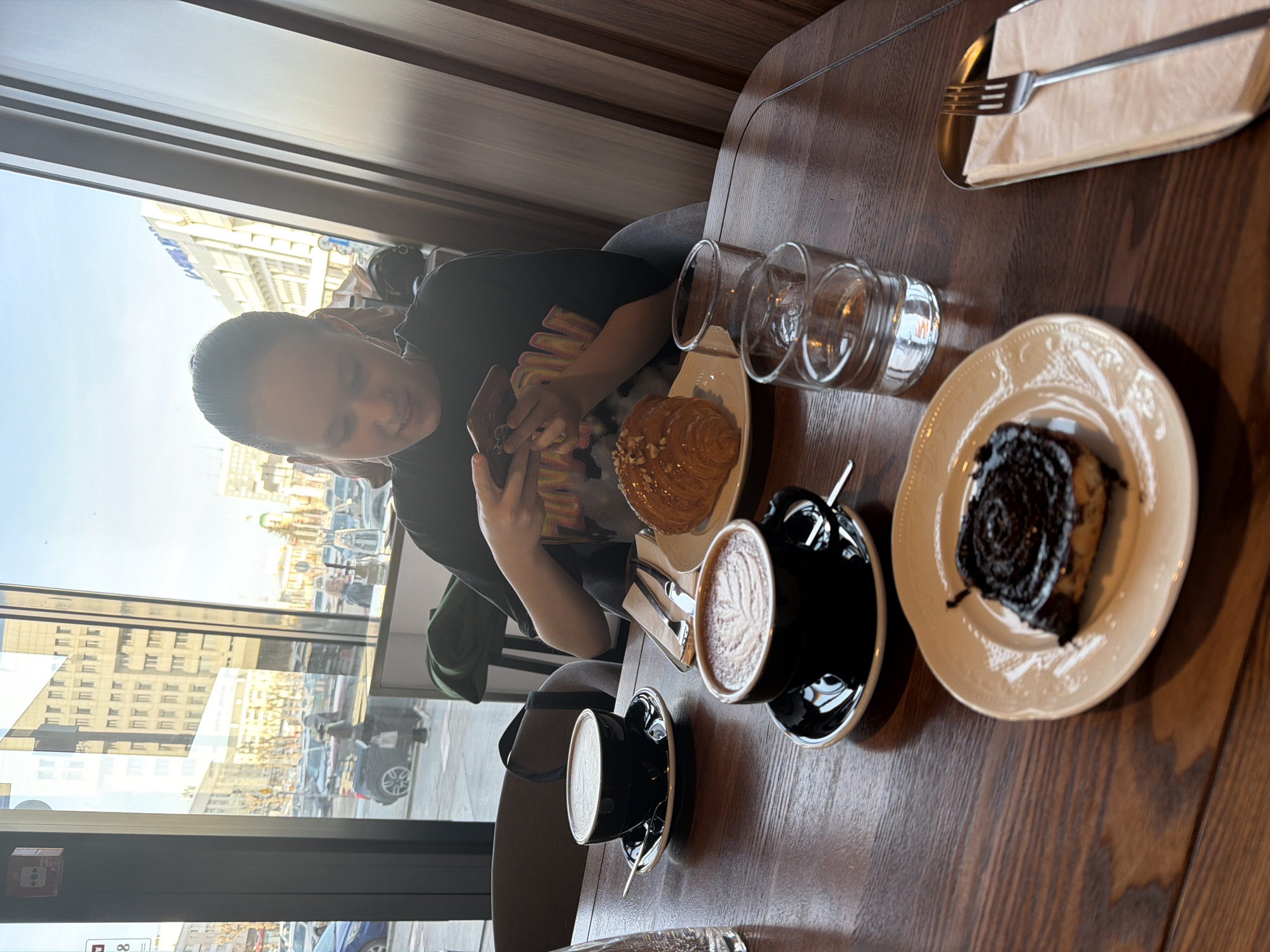 The image captures an indoor scene at a café in Perm, Russia. A person with short dark hair sits near a window looking at their phone. Outside the window is a view of the city, including building facades and street traffic. On the table, there are two black cups of coffee on saucers with spoons, a croissant, a piece of cake on a plate, two clear glasses stacked together, and a fork on top of a napkin. The table is made of dark brown wood, and a vertical wooden paneling appears in the background. The scene is well-lit, likely due to the natural light coming through the window.