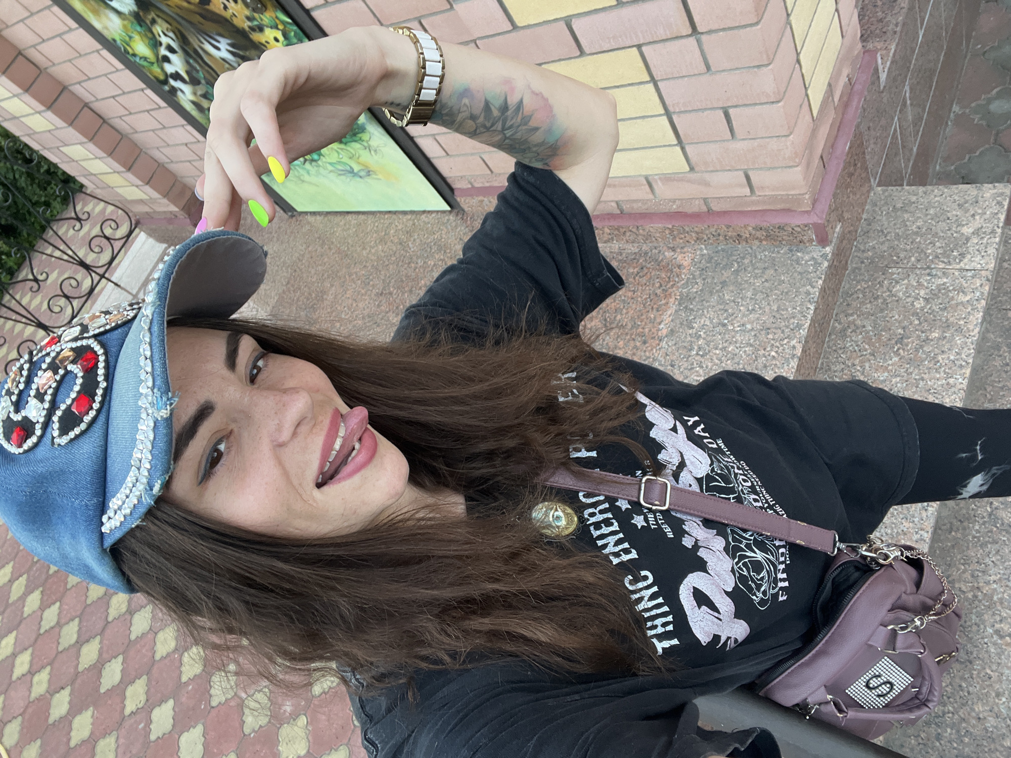 A woman with dark hair and a blue denim cap with rhinestones and red embellishments is sticking her tongue out and touching her cap. She is wearing a black t-shirt with a graphic print and a purple crossbody bag. She has neon green and yellow nail polish on her fingers. A watch with a white band is visible on her right wrist. She has a colorful tattoo on her right forearm. The background appears to be an outdoor setting with brickwork and a fence. There is a painting on the wall to the right of the woman. The paving stones on the ground are red and beige. This image was taken in Krasnoe-na-Volge, Russia.