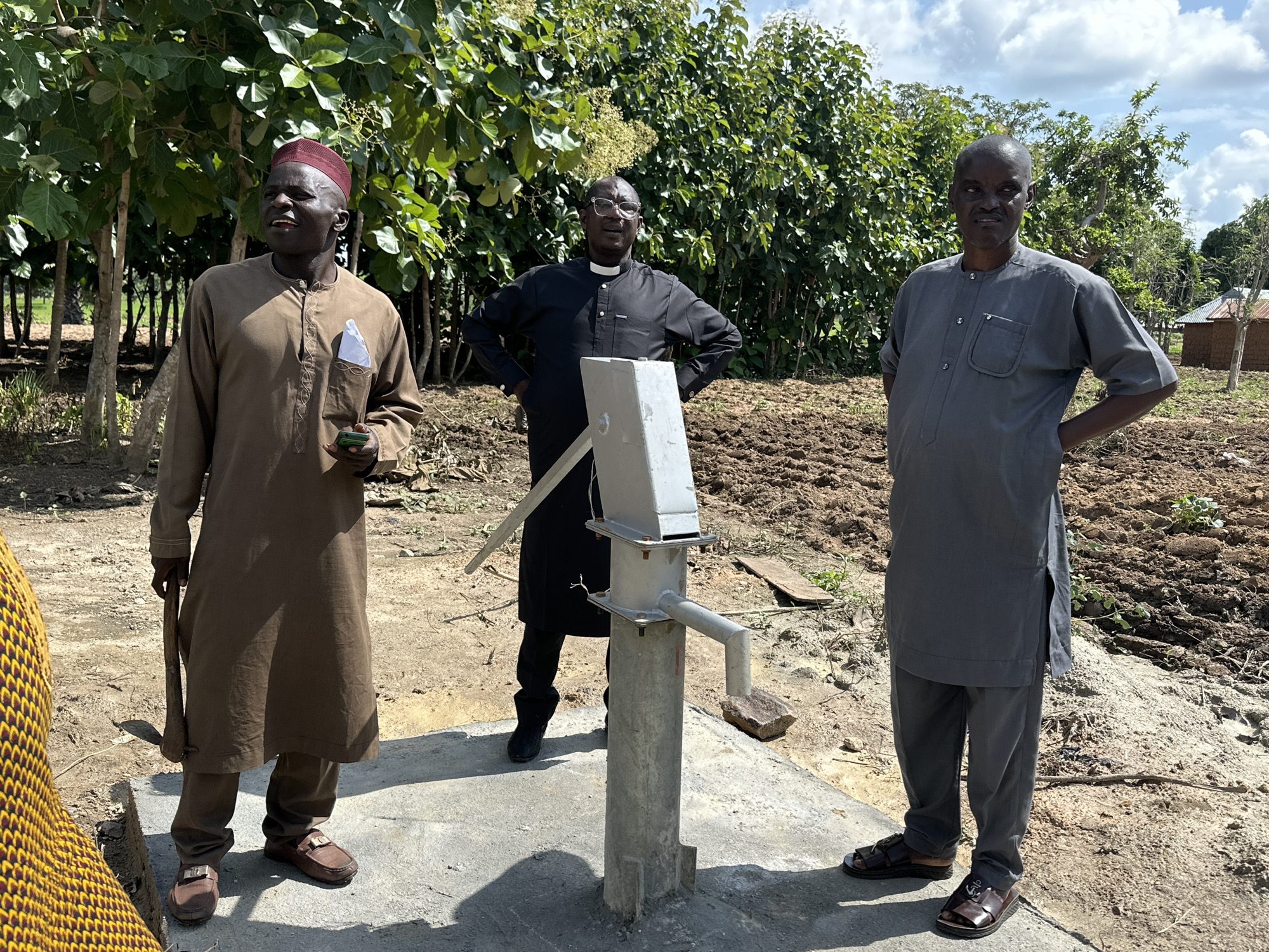 The image depicts three men standing outdoors in Afogo, Nigeria, near a hand pump. The man on the left is wearing a brown tunic and pants, a red cap, and brown loafers. He is holding a small green object and a tool that resembles a hoe. The man in the center, dressed in a black clerical shirt with a white collar, glasses, and black pants, stands with his hands on his hips. The man on the right is wearing a grey tunic and pants and brown sandals, also with his hands on his hips. They are positioned on a concrete pad surrounding the grey metal hand pump, which is situated in a dirt field with some vegetation and scattered small buildings in the background. The weather appears to be clear with a partly cloudy sky.