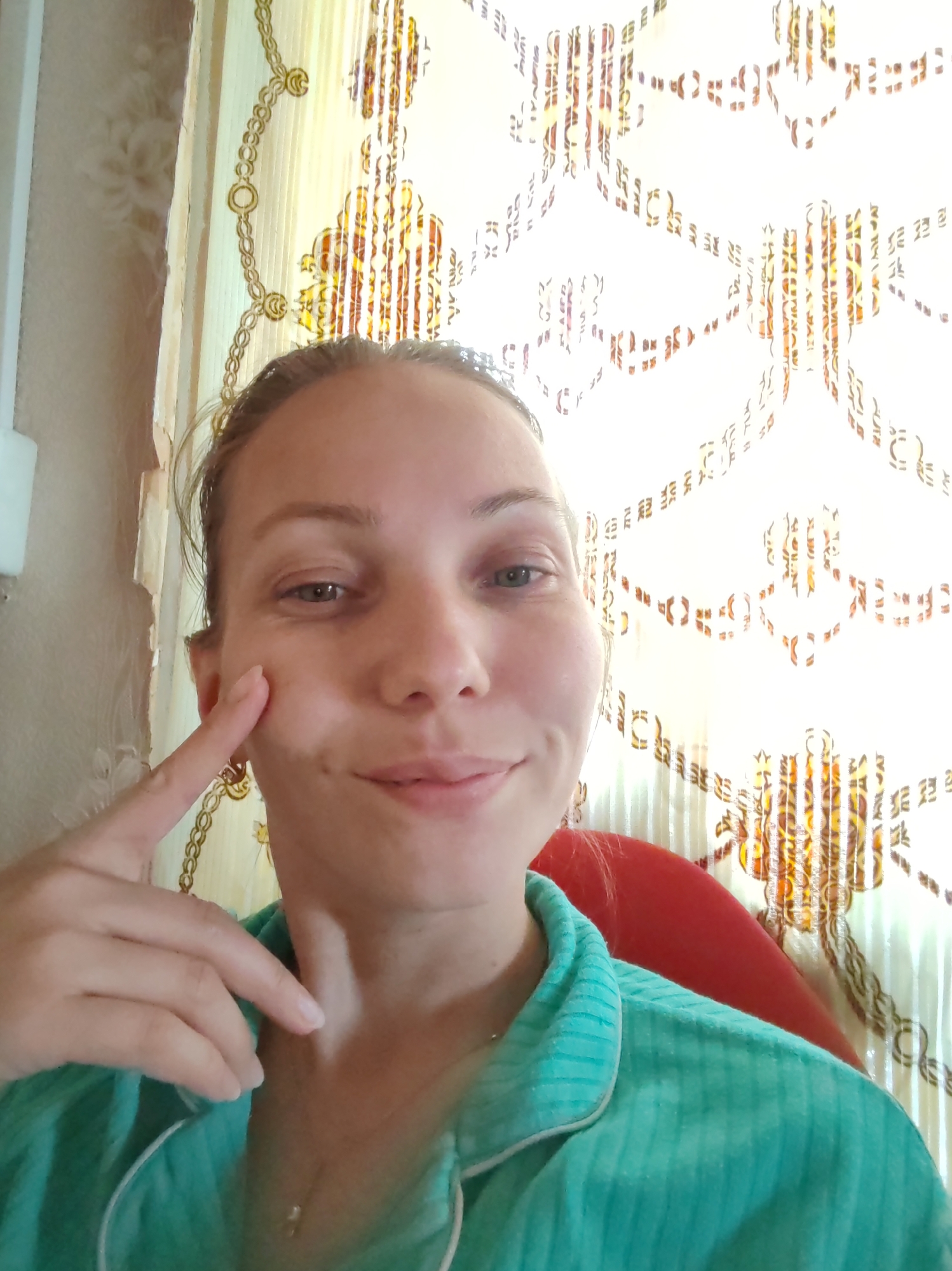 In a close-up, selfie-style shot, a fair-skinned woman with light brown hair pulled back is looking directly at the camera. Her left hand is raised, with her index finger touching her cheek. She is wearing a teal-colored pajama top with white trim. Behind her, a patterned curtain with a gold chain motif is visible, and to her left, a portion of a wall with peeling wallpaper can be seen. The overall lighting is bright, suggesting it's daytime and possibly near a window. No specific location in Чухлэм, Russia, can be determined from the visual elements presented.